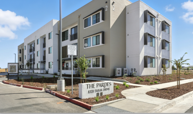 Exterior view of a modern apartment building with a landscaped sidewalk and a sign reading "The Pardes, 8355 Tarak Drive" in front.