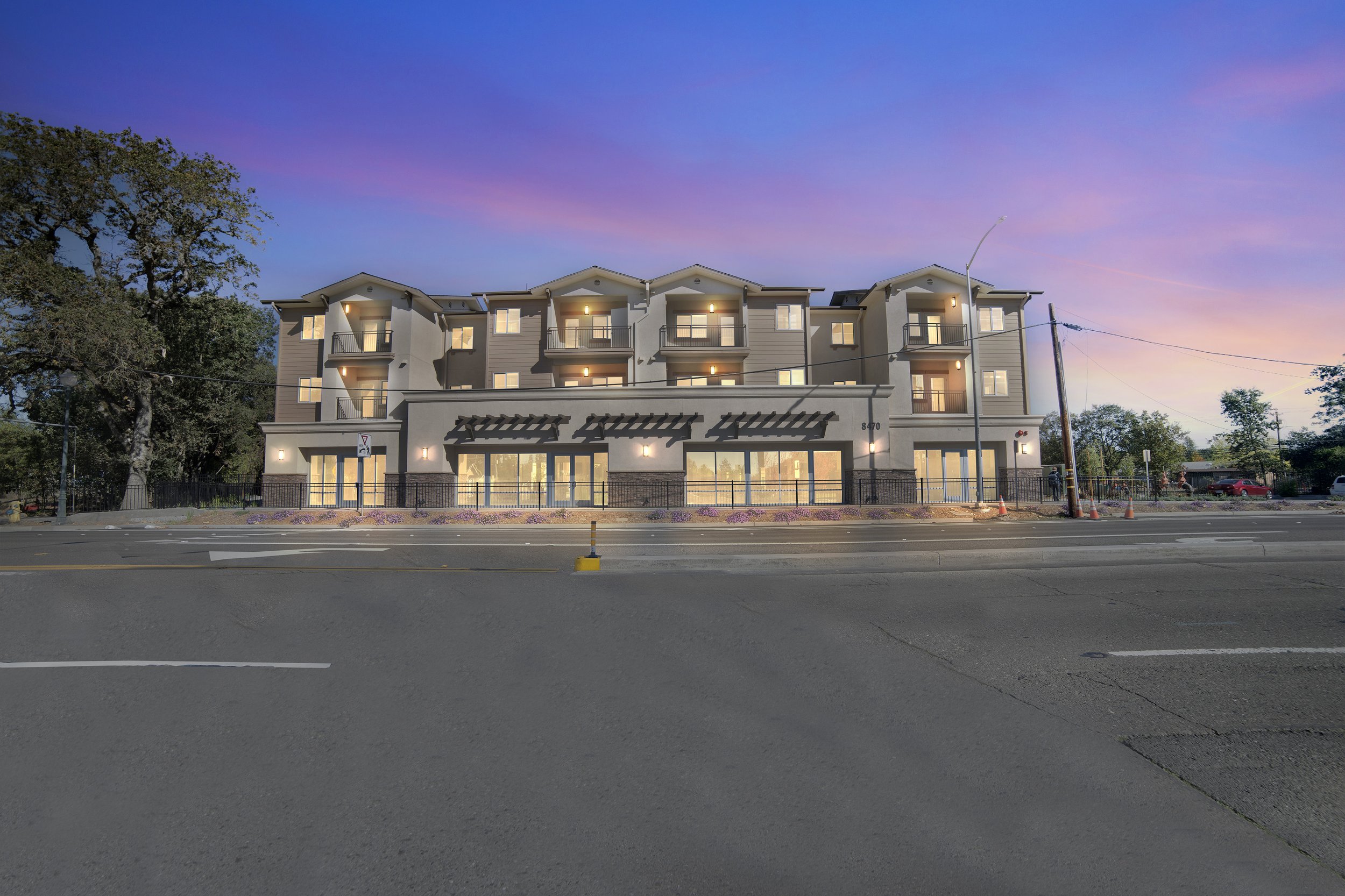 The image shows a modern multi-story apartment building with illuminated windows and balconies at dusk, with a colorful sky in the background. There are trees on the left and right sides, and a street with traffic cones and vehicles in the foreground.
