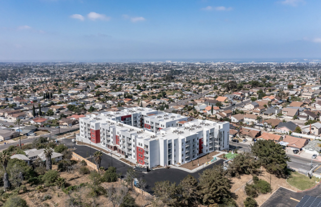 Aerial view of a modern white apartment building with red accents surrounded by a residential neighborhood with single-family homes in a cityscape.