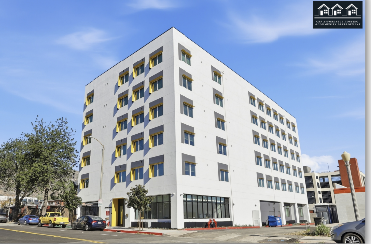 Modern white multi-story apartment building with yellow window awnings, situated on a city street with parked cars and trees, under a blue sky.