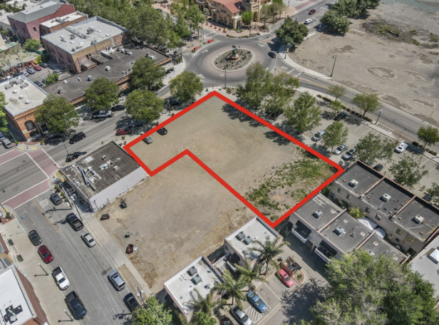 A bird's eye view of an urban area with a vacant lot outlined in red, surrounded by trees, buildings, and parking lots, with a roundabout featuring a statue in the background.