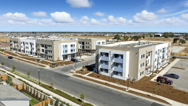 Residential apartment complex with multiple buildings, parking lots, and landscaped walkways under a partly cloudy sky.