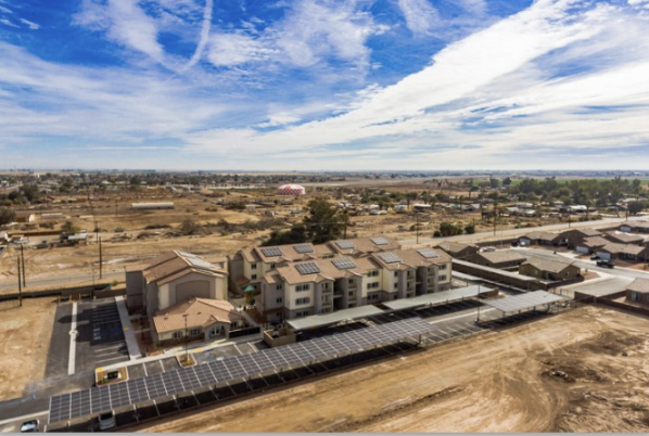 A solar panel installation in front of a multi-family residential building in an arid area under a partly cloudy sky.