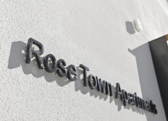 Close-up of a sign on a white wall that reads 'Rose Tow  Apartments' with shadows cast by the letters.