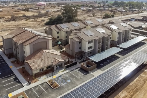 Aerial view of a residential complex with multiple townhouses and parking lot, adjacent to a building with solar panels on the roof in a dry area.