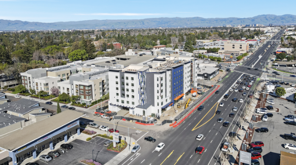 Aerial view of a busy city street with cars, buildings, and storefronts, with mountains in the background.