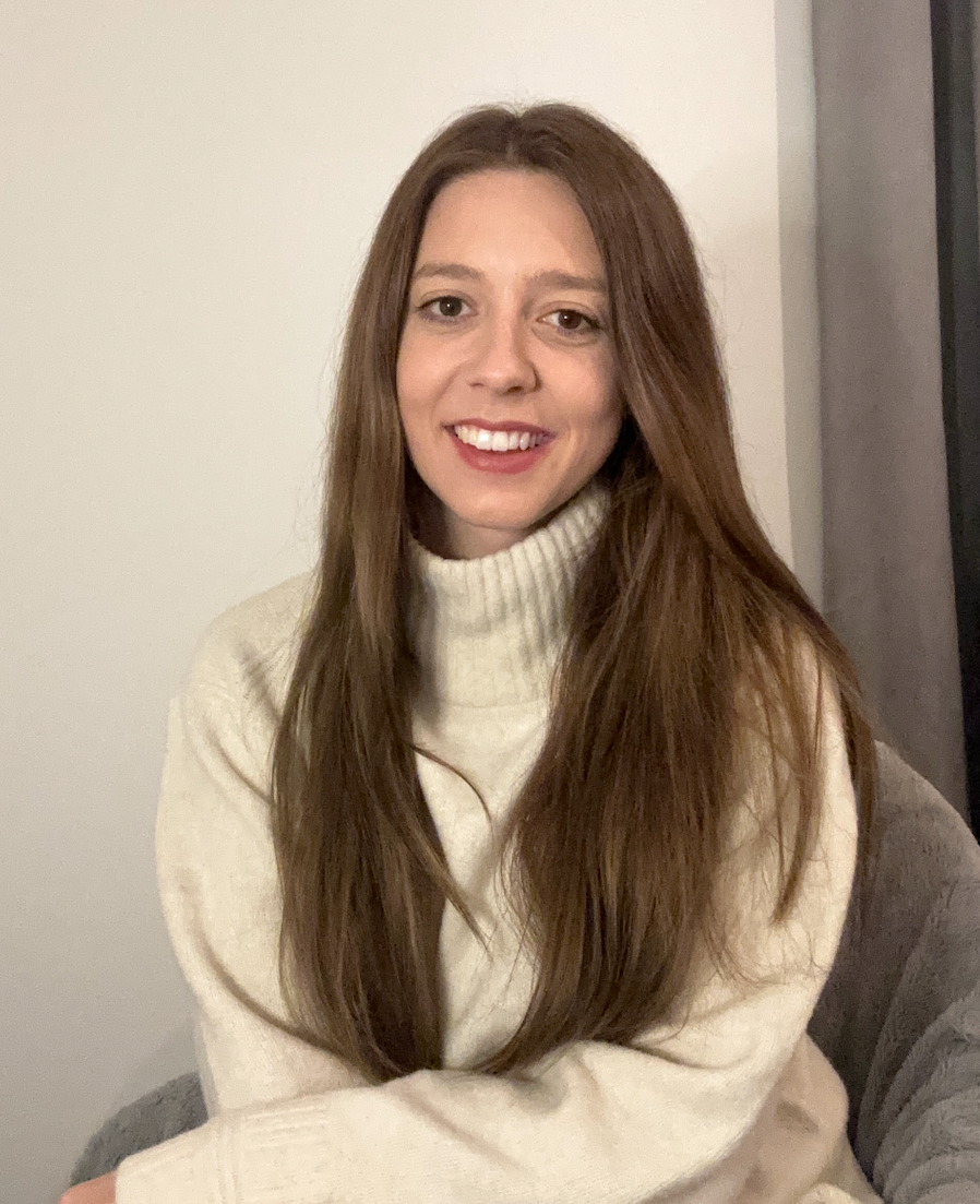 A young woman with long brown hair, smiling, wearing a beige turtleneck sweater, sitting indoors against a plain wall.