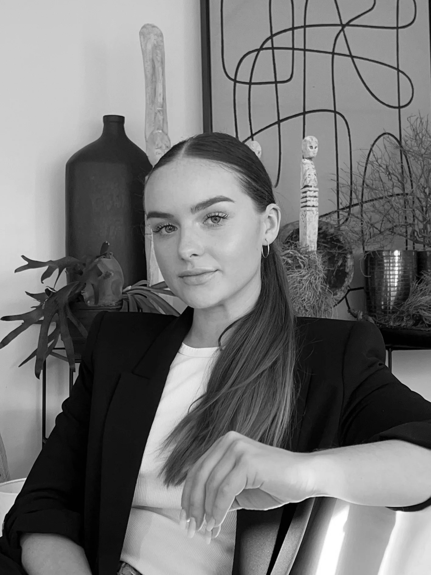 A woman with long straight hair, wearing a black blazer and white top, sitting in a room with decorative objects, plants, and abstract art in the background.
