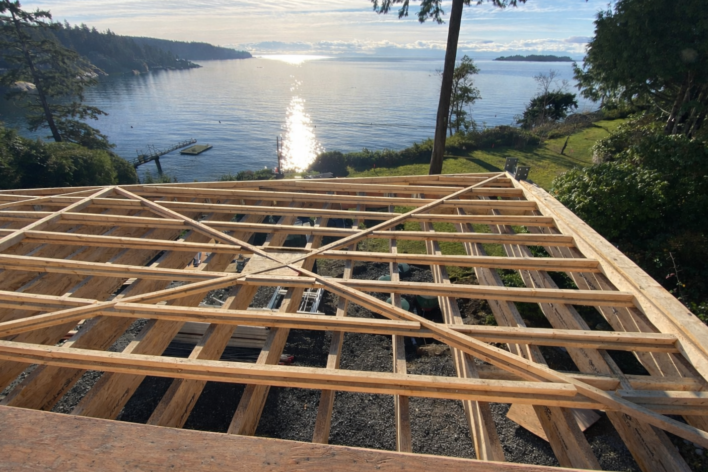 Wooden framing structure for a building or deck overlooking a body of water with a dock and trees in the background.