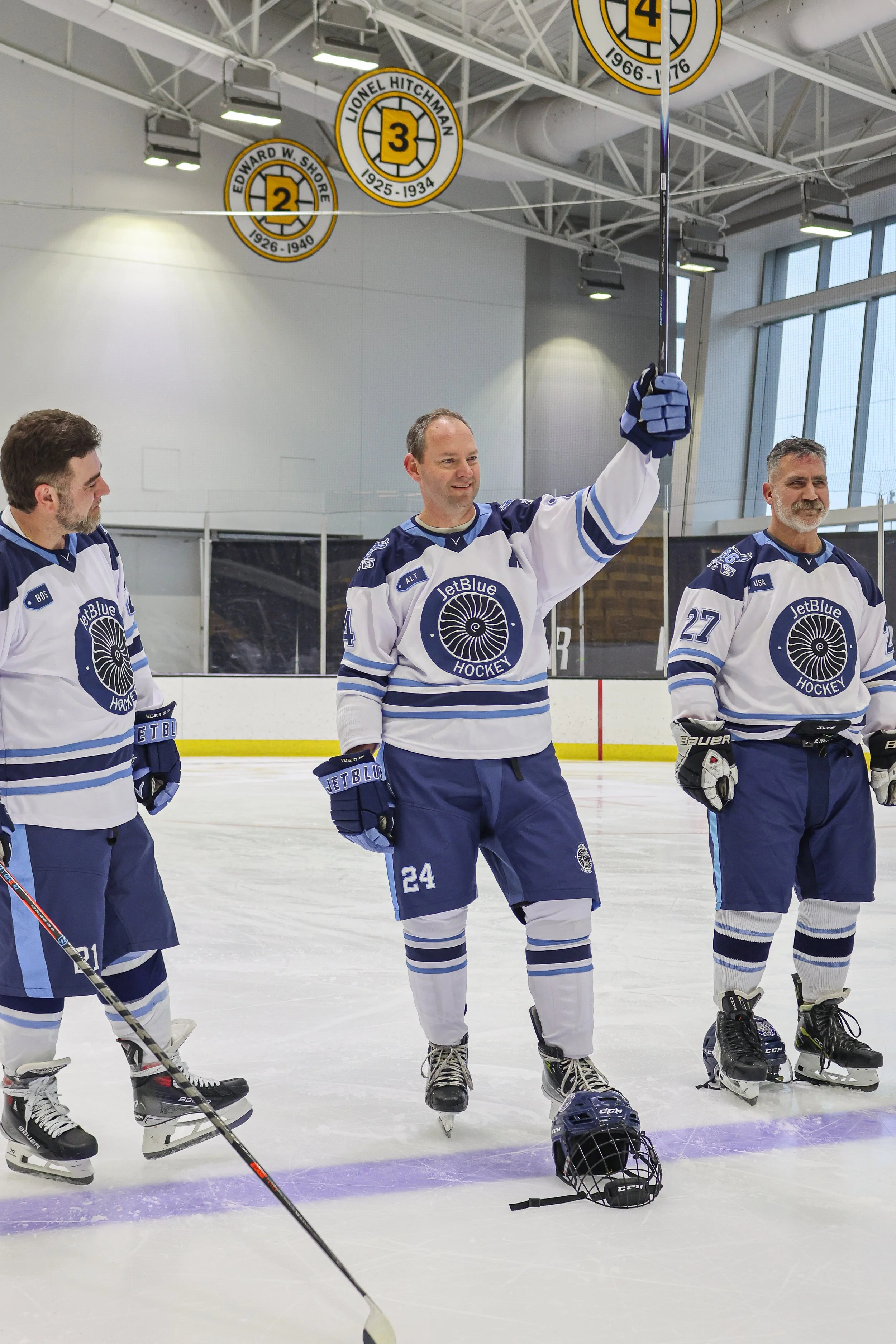 Three ice hockey players in white and blue uniforms standing on ice rink with one holding a hockey stick aloft.
