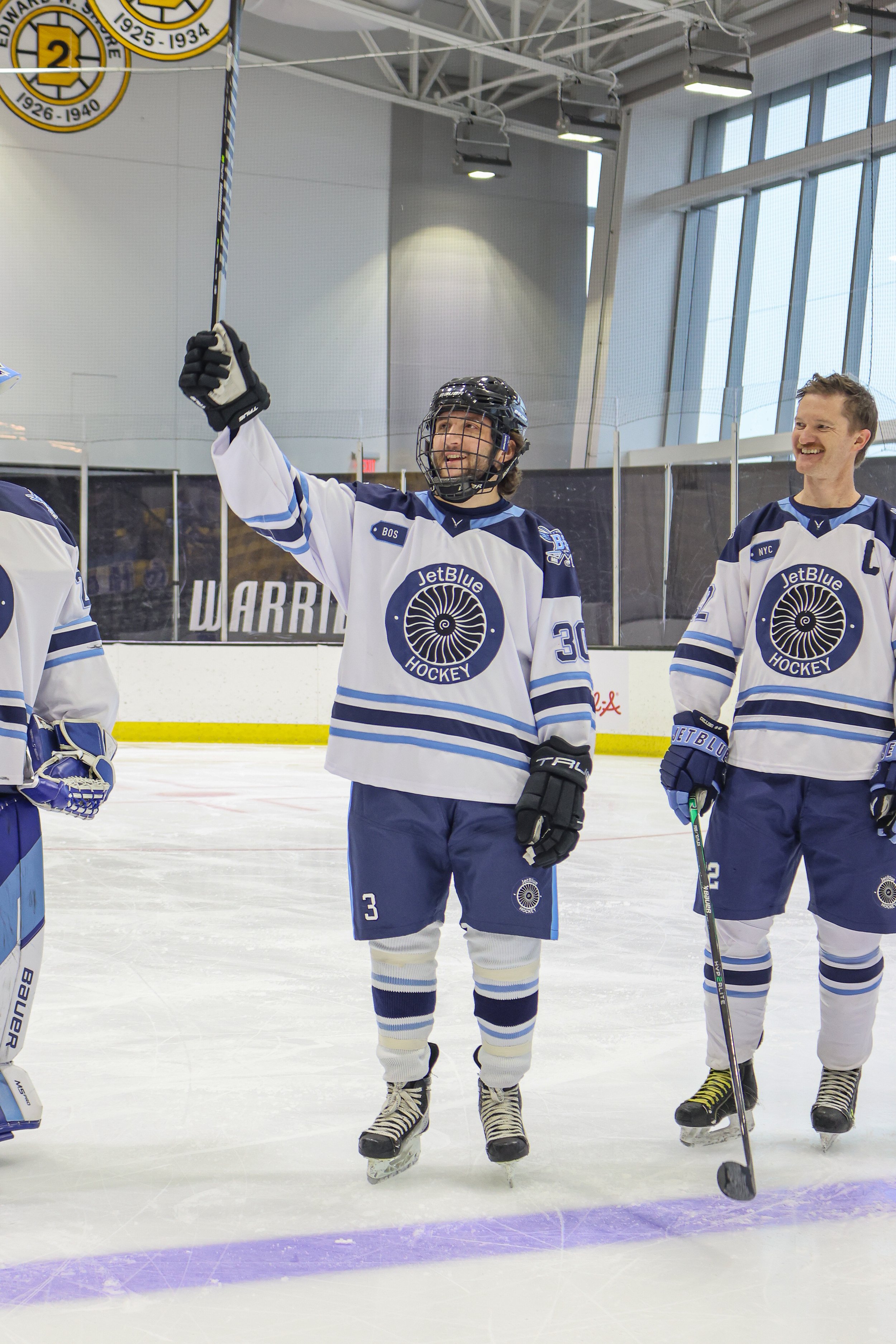 Hockey players in white and blue jerseys celebrating on the ice rink. One player is raising his arm, smiling, and wearing a helmet and gloves. Others nearby are also smiling and holding hockey sticks.