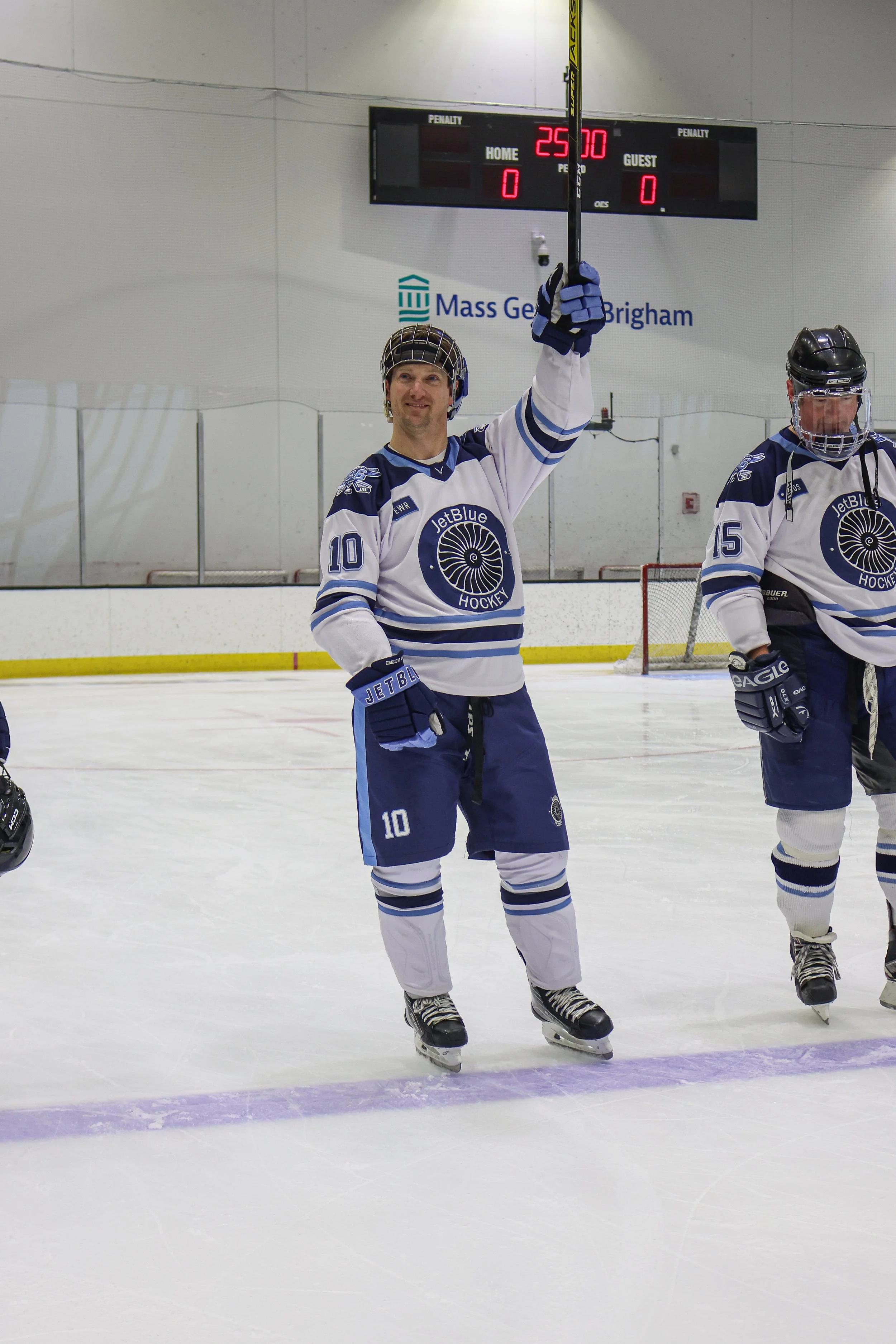 A male hockey player in a blue and white jersey with the number 10, holding a hockey stick upright over his head after a game, standing on an ice rink.