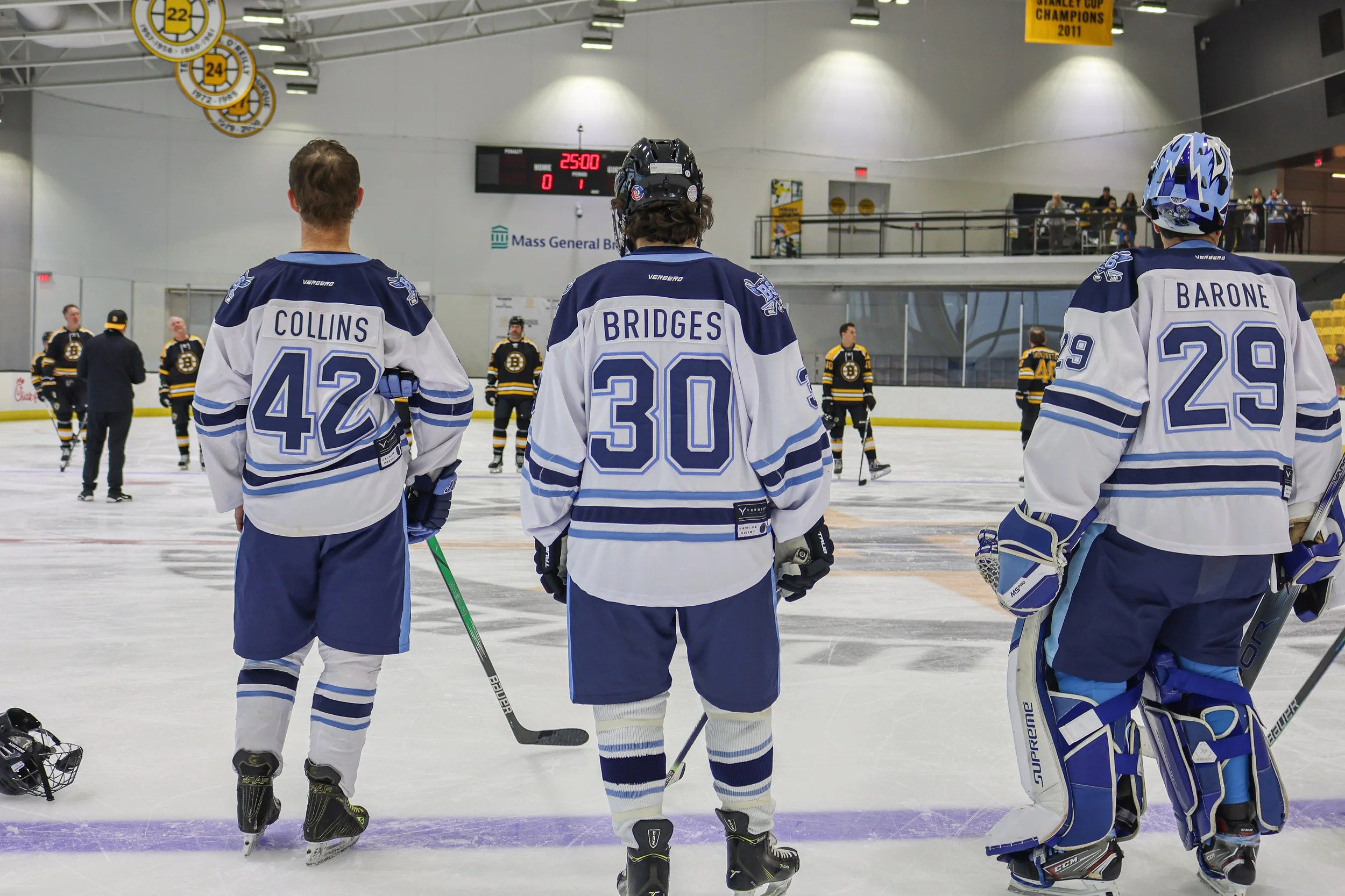 Three hockey players in white and blue jerseys standing on ice rink facing away, with more players in the background, and a scoreboard showing 25:00 minutes.