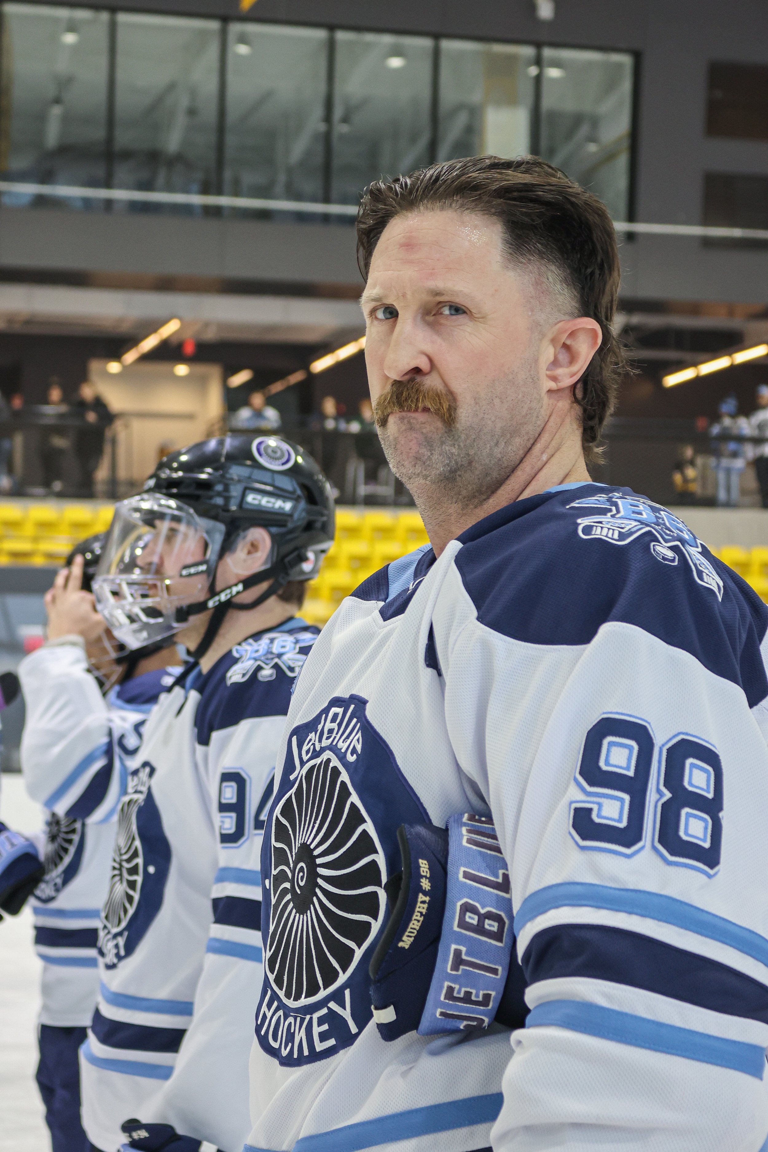A hockey player with a mustache and beard wearing a white and blue uniform with the number 98, standing on the ice rink with other players and spectators in the background.