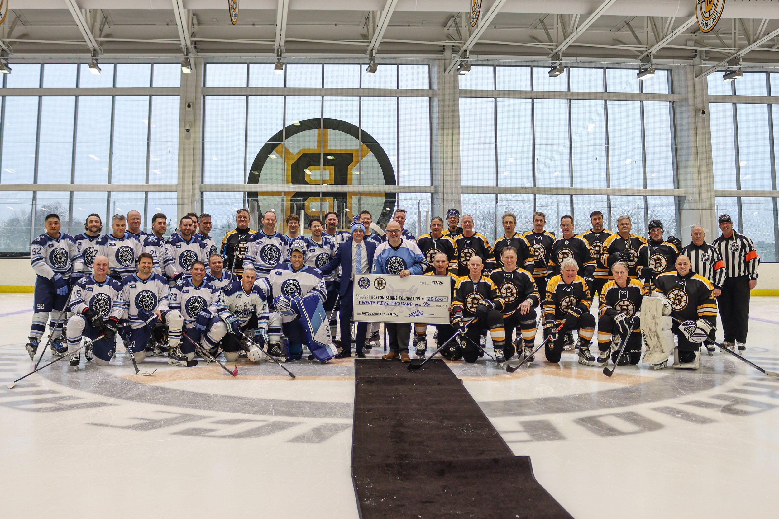 Group of hockey players, coaches, and officials on an ice rink holding a large check for $25,000, celebrating fundraising success, with team logos and a large logo in the background.