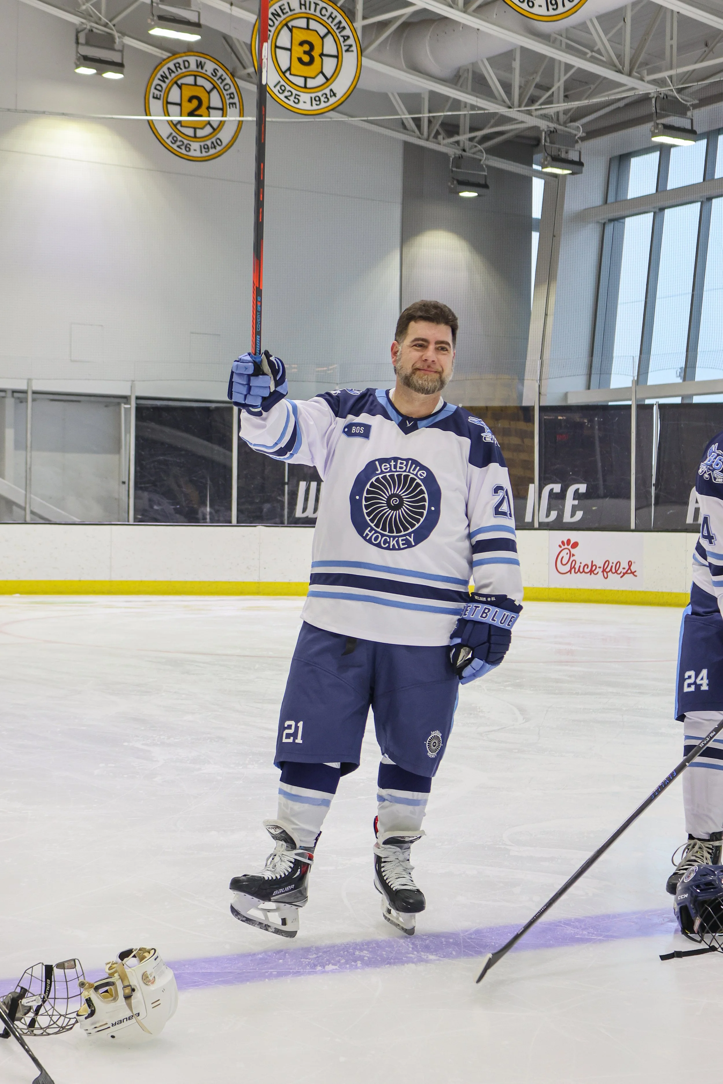 A male hockey player in a blue and white jersey hanging on an indoor ice rink. He is holding a hockey stick up in the air and wearing ice skates. The helmet is placed on the ice near his feet.