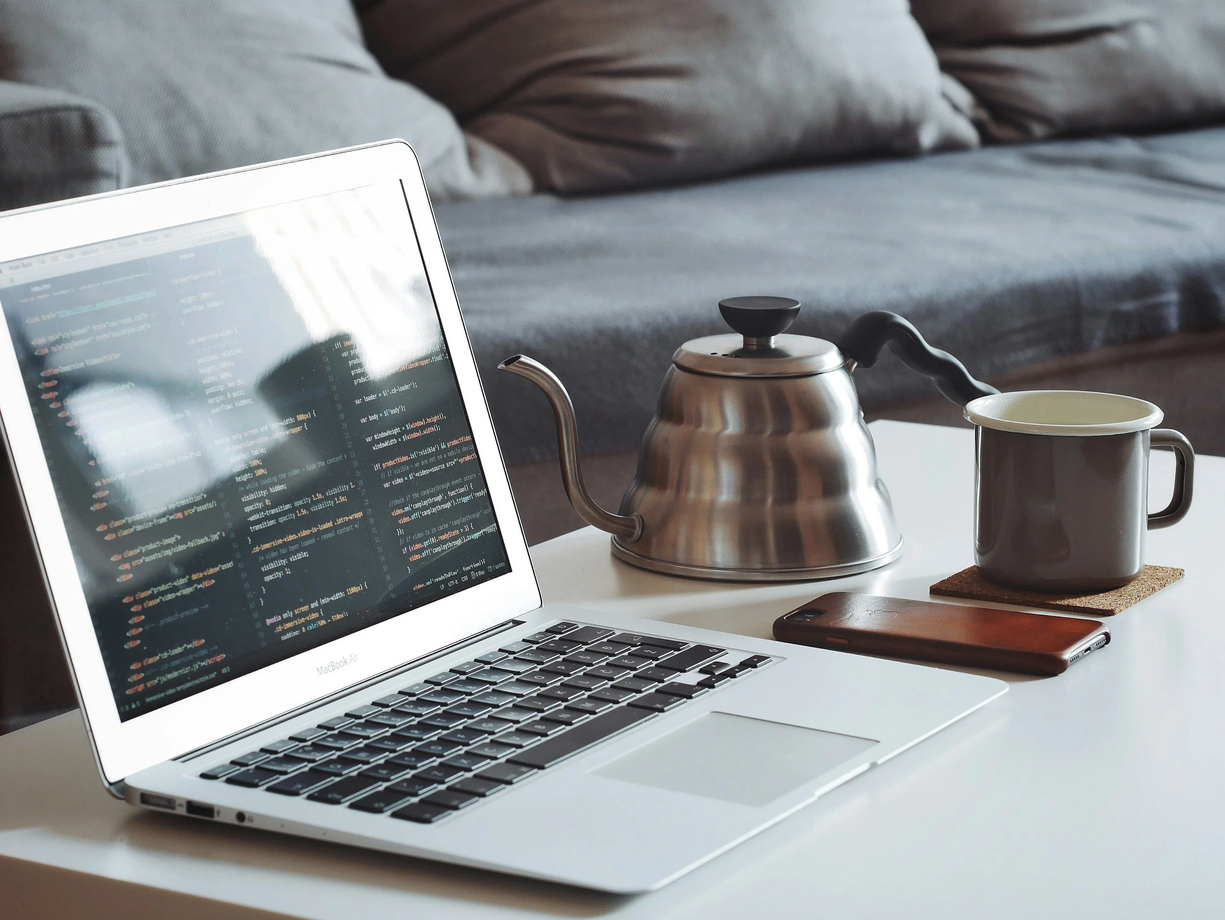 Laptop with code on screen, metal teapot, gray coffee mug on cork coaster, smartphone, and gray couch in background.