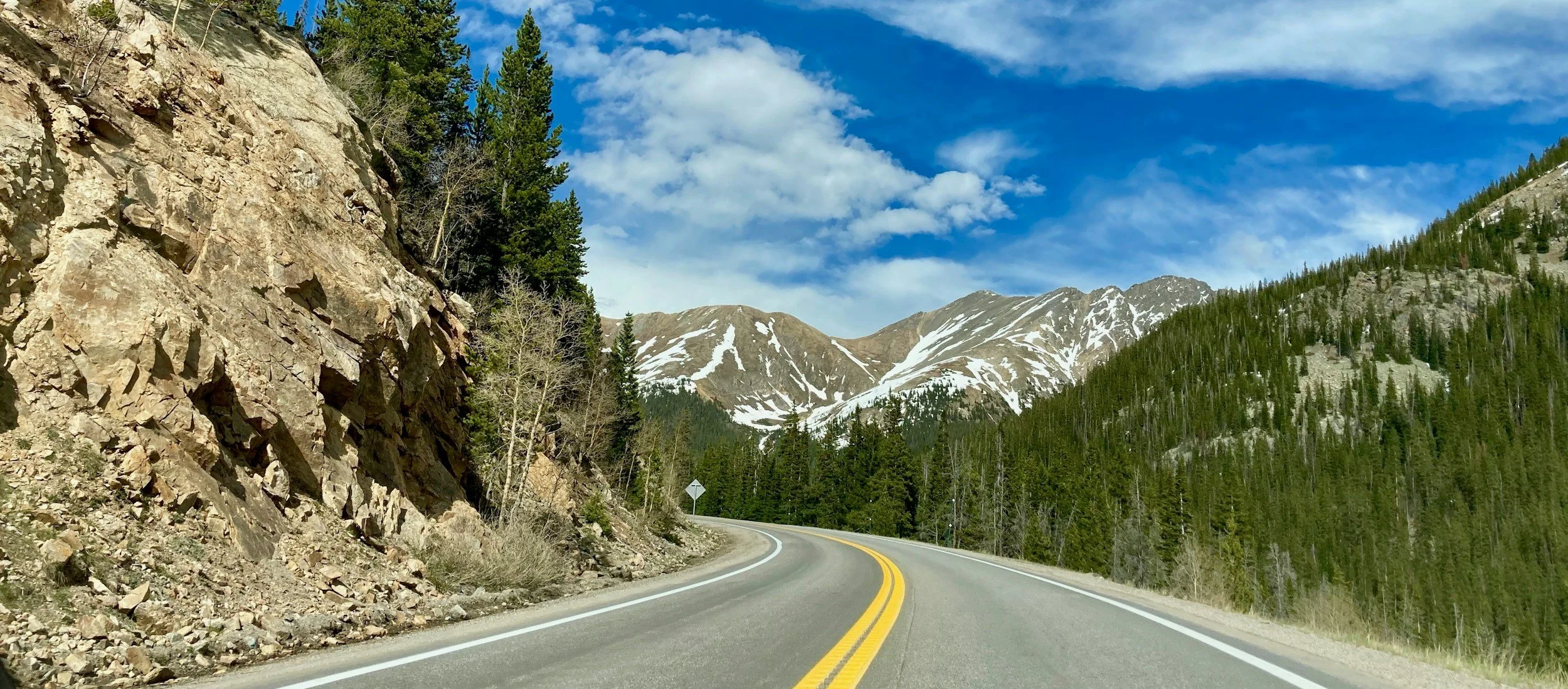 Curving mountain road with yellow lines, surrounded by pine trees and rocky slopes, snow-capped mountains in the background under a blue sky with scattered clouds.