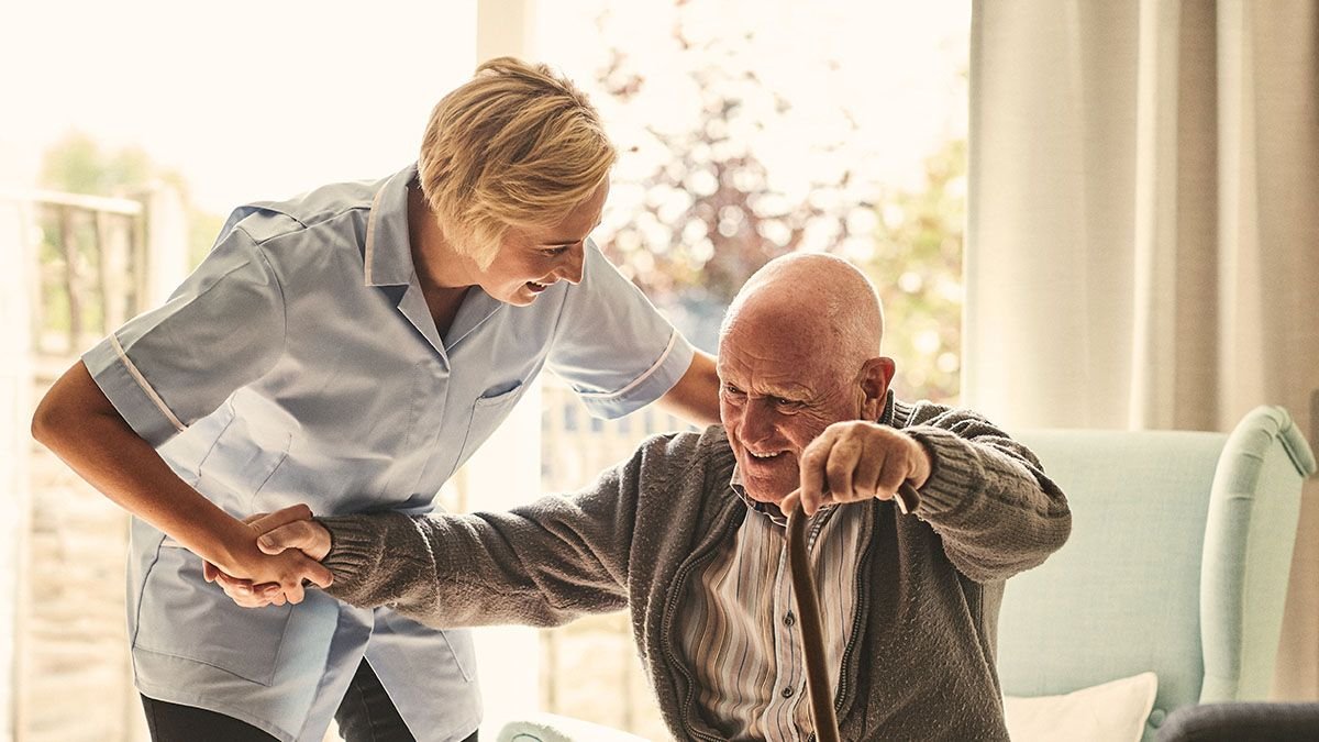 A young woman healthcare worker helping an elderly man with a cane, smiling indoors near a window.