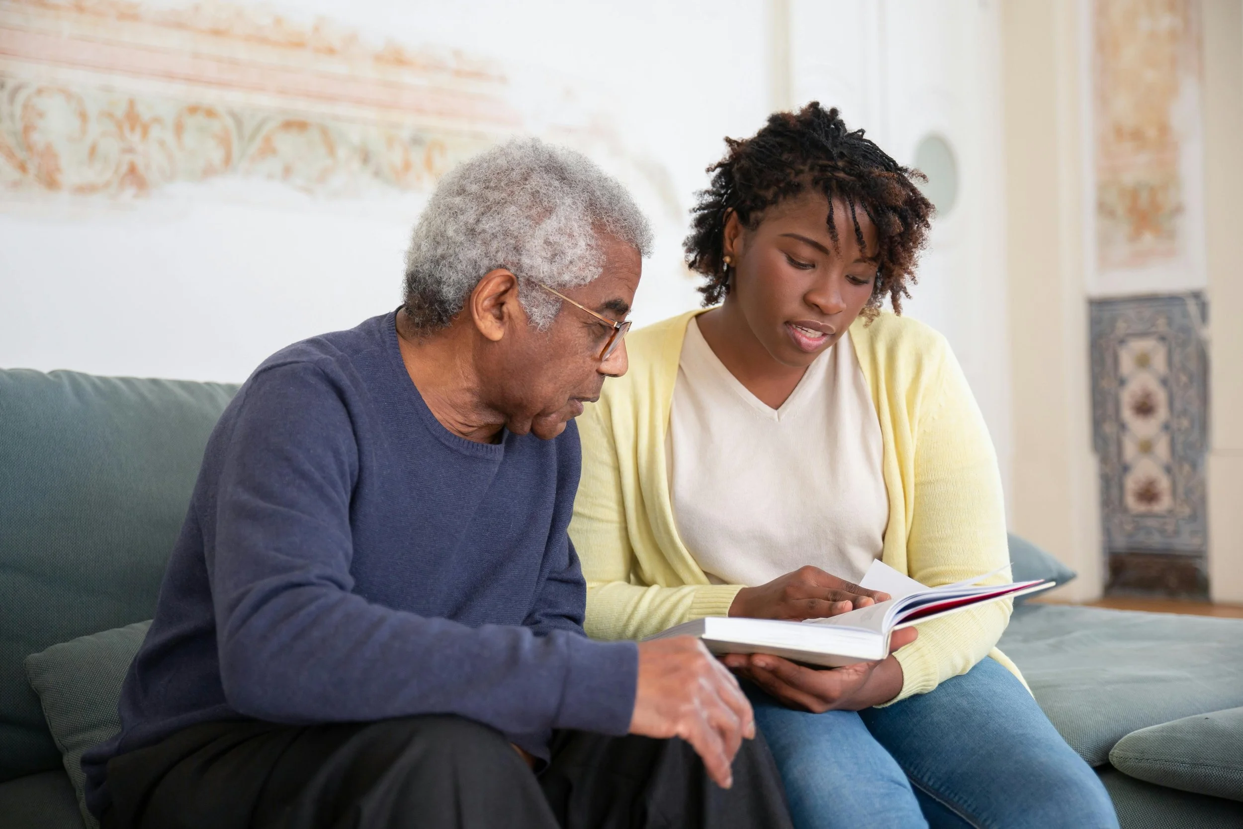 An elderly man with gray hair and glasses sitting on a green couch next to a young woman with dark curly hair, both looking at an open book together.