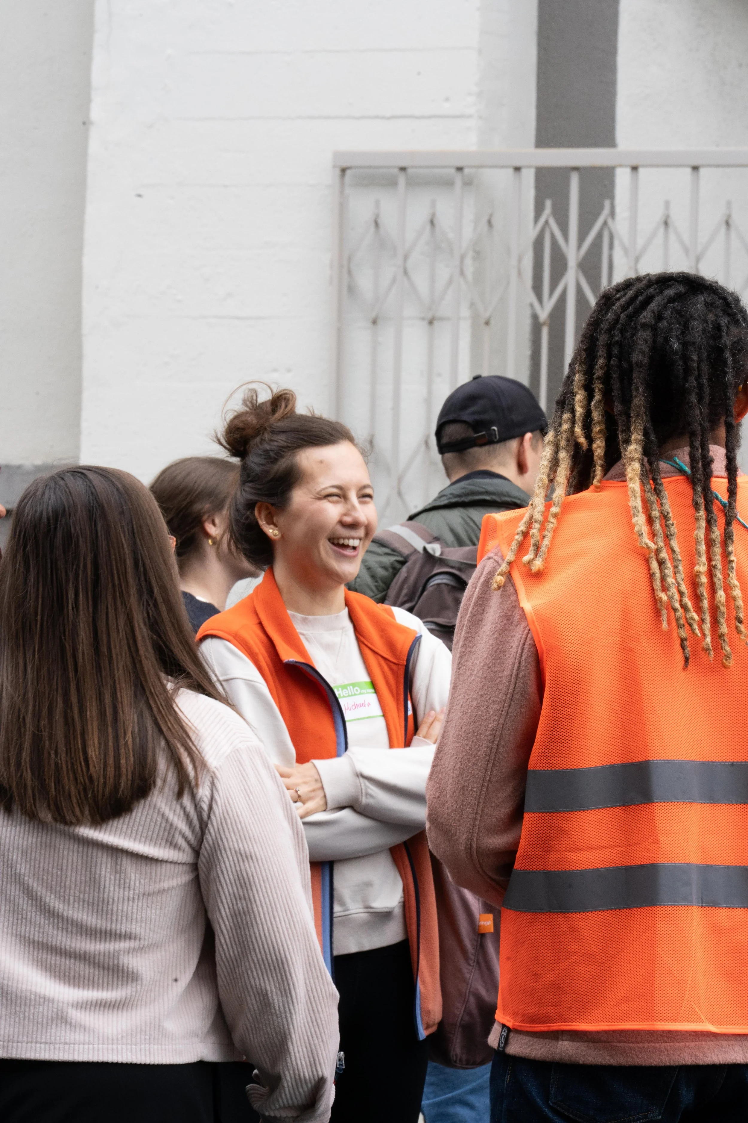Group of people engaging in conversation, with woman smiling, wearing orange vest, in a casual indoor setting.