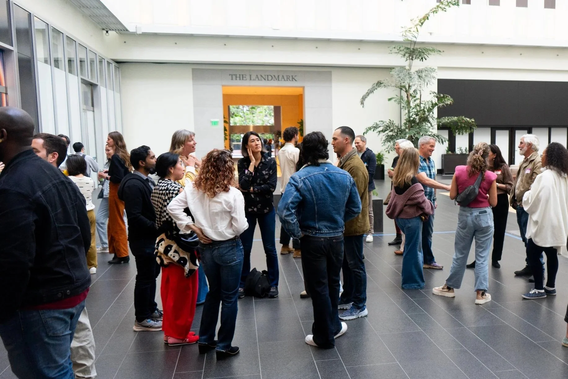 A diverse group of people socializing inside a modern building with large windows and decorative plants, under a sign that reads 'The Landmark.'