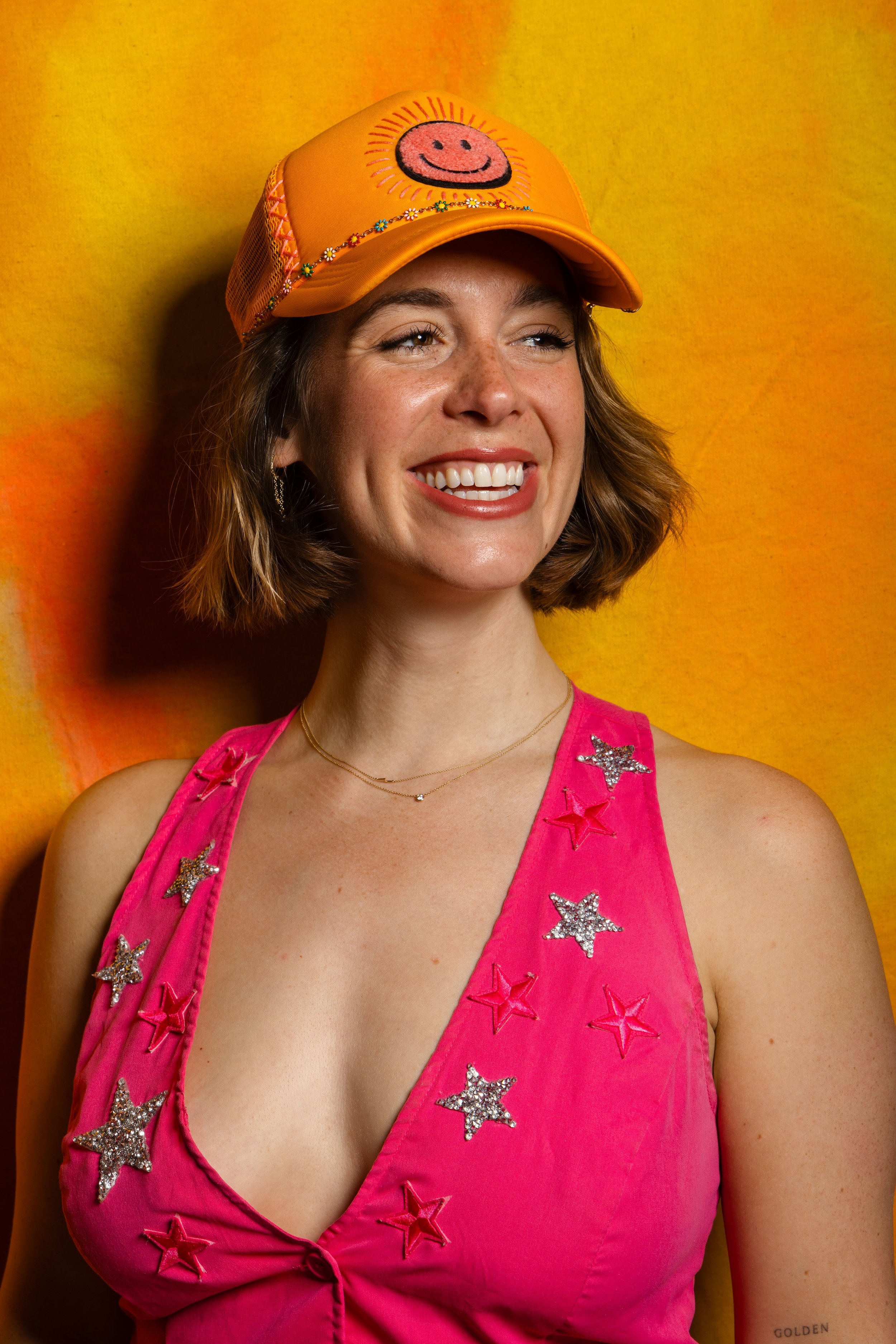 A woman with short wavy hair wearing a pink top decorated with sequined star patches, a gold necklace, and an orange baseball cap with a smiling face design, standing in front of a yellow-orange background.