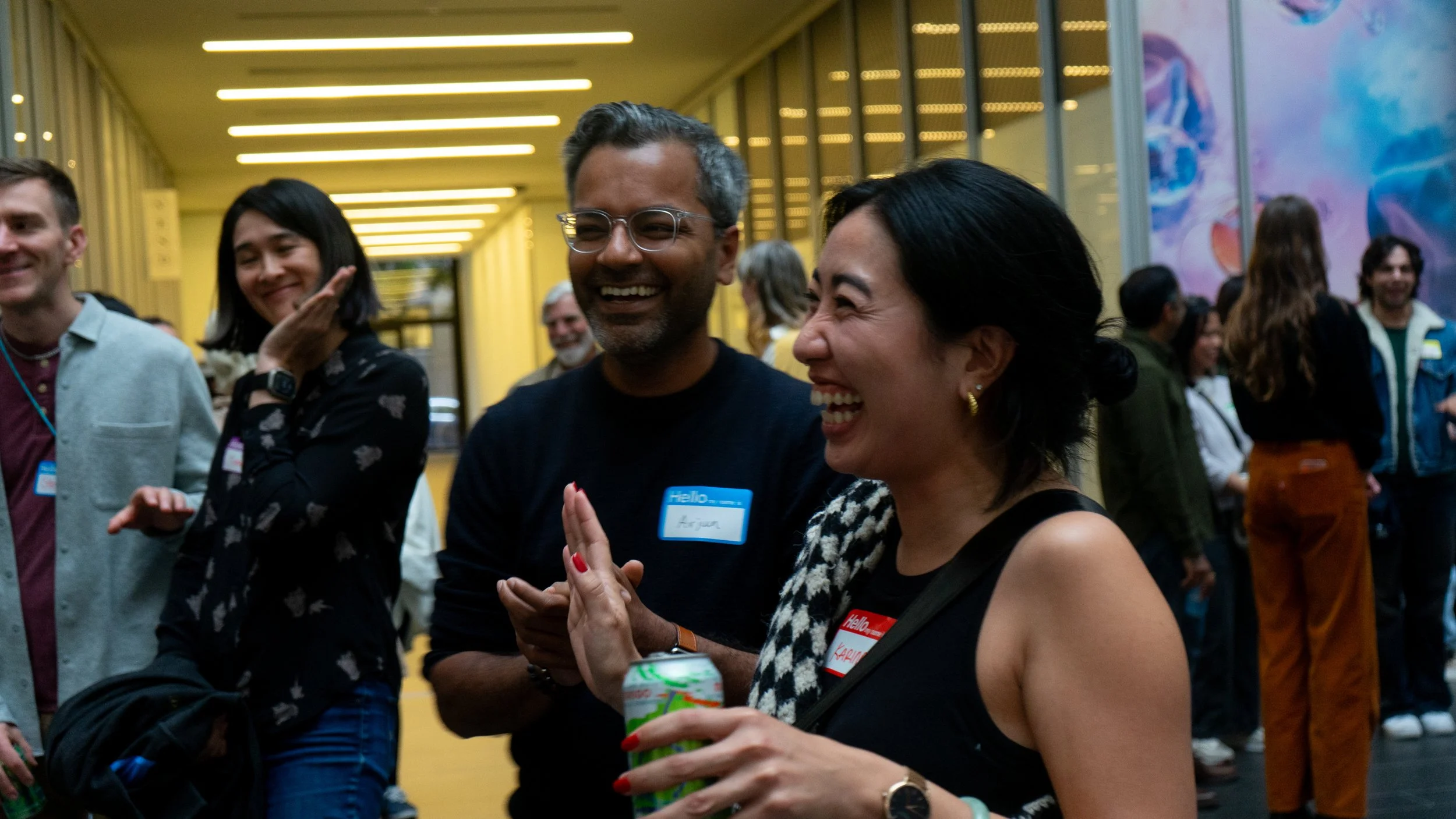 A group of people at a social event, engaging in conversation and laughing inside a bright, modern space with yellow walls and glass panels.