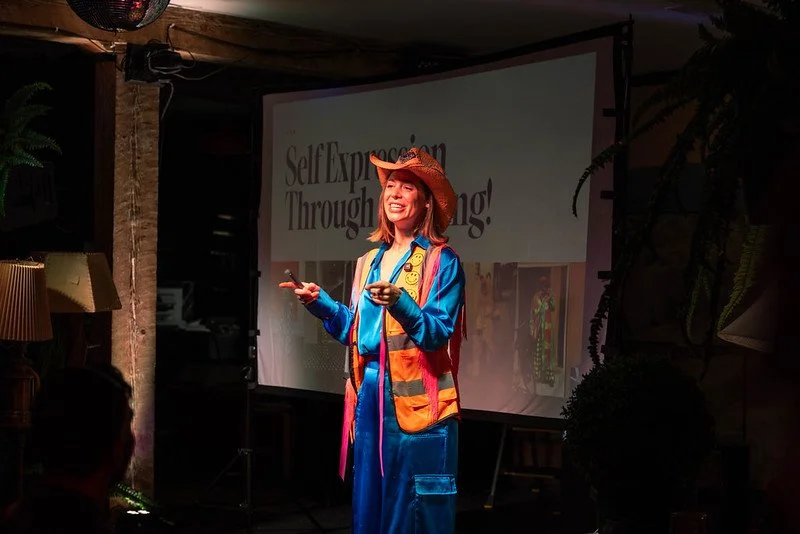A woman dressed in colorful adventure attire, wearing a straw hat, giving a presentation on a stage with a projection screen behind her that reads 'Self-Expression Through ...'.