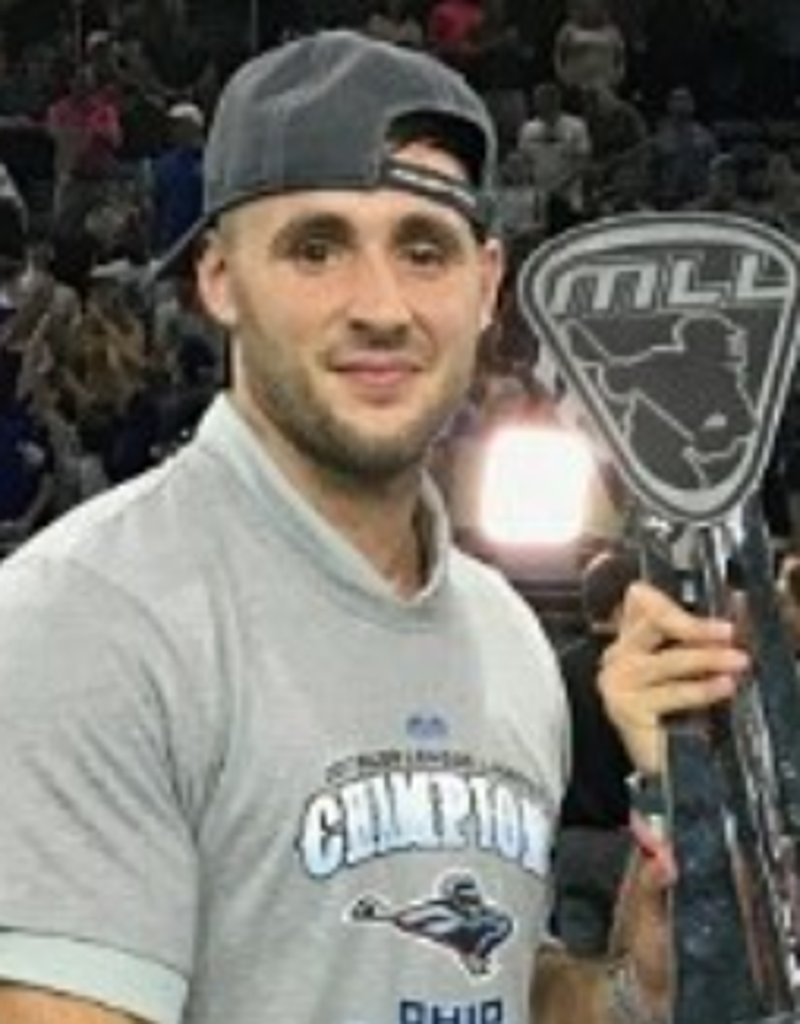 Young man wearing a gray T-shirt with 'Champions' and a sports team logo, a backward snapback cap, and holding a hockey stick, in a crowded arena.