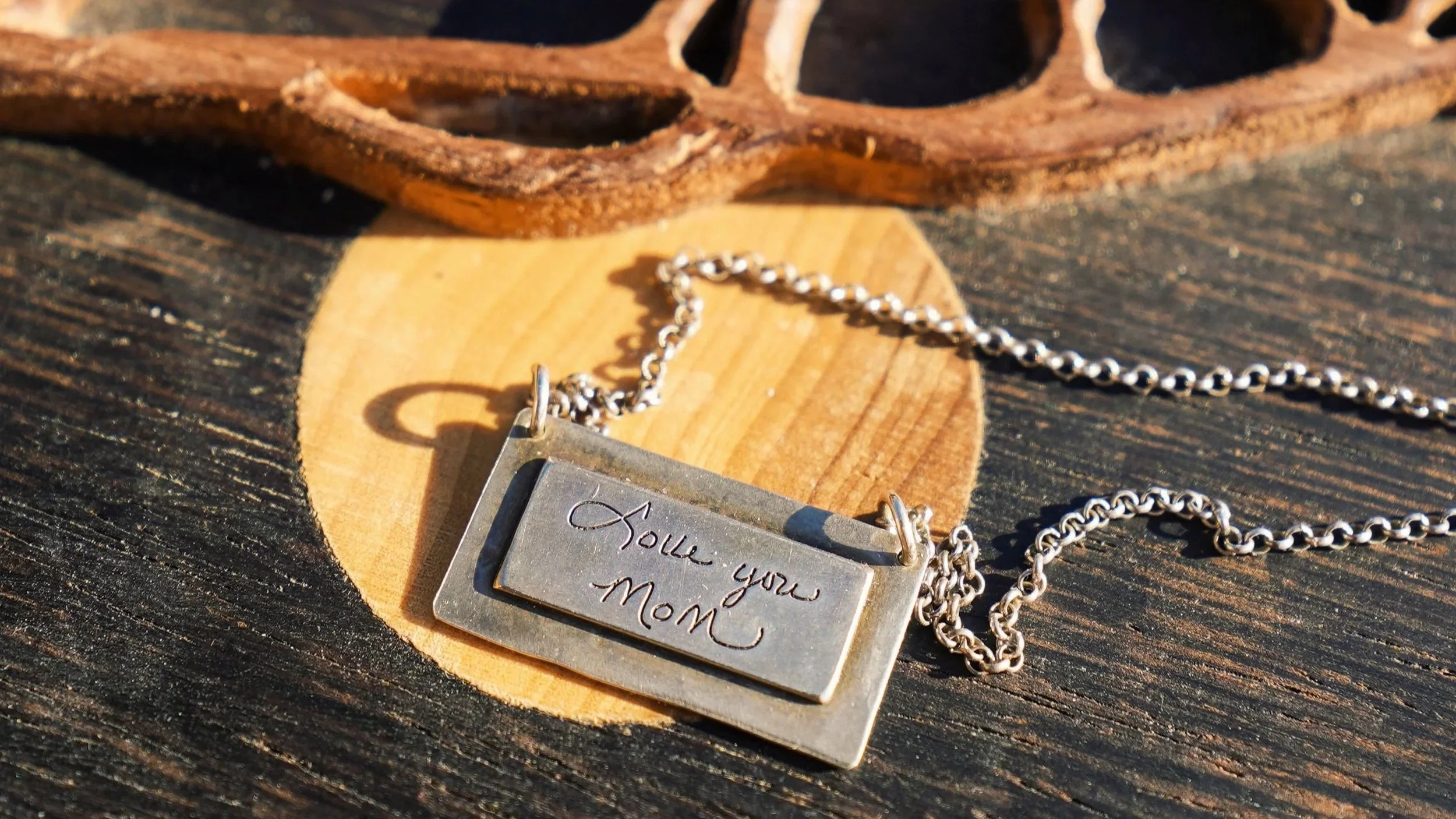 A silver necklace with a rectangular pendant engraved with a message, placed on a wooden surface with a carved wooden object in the background.