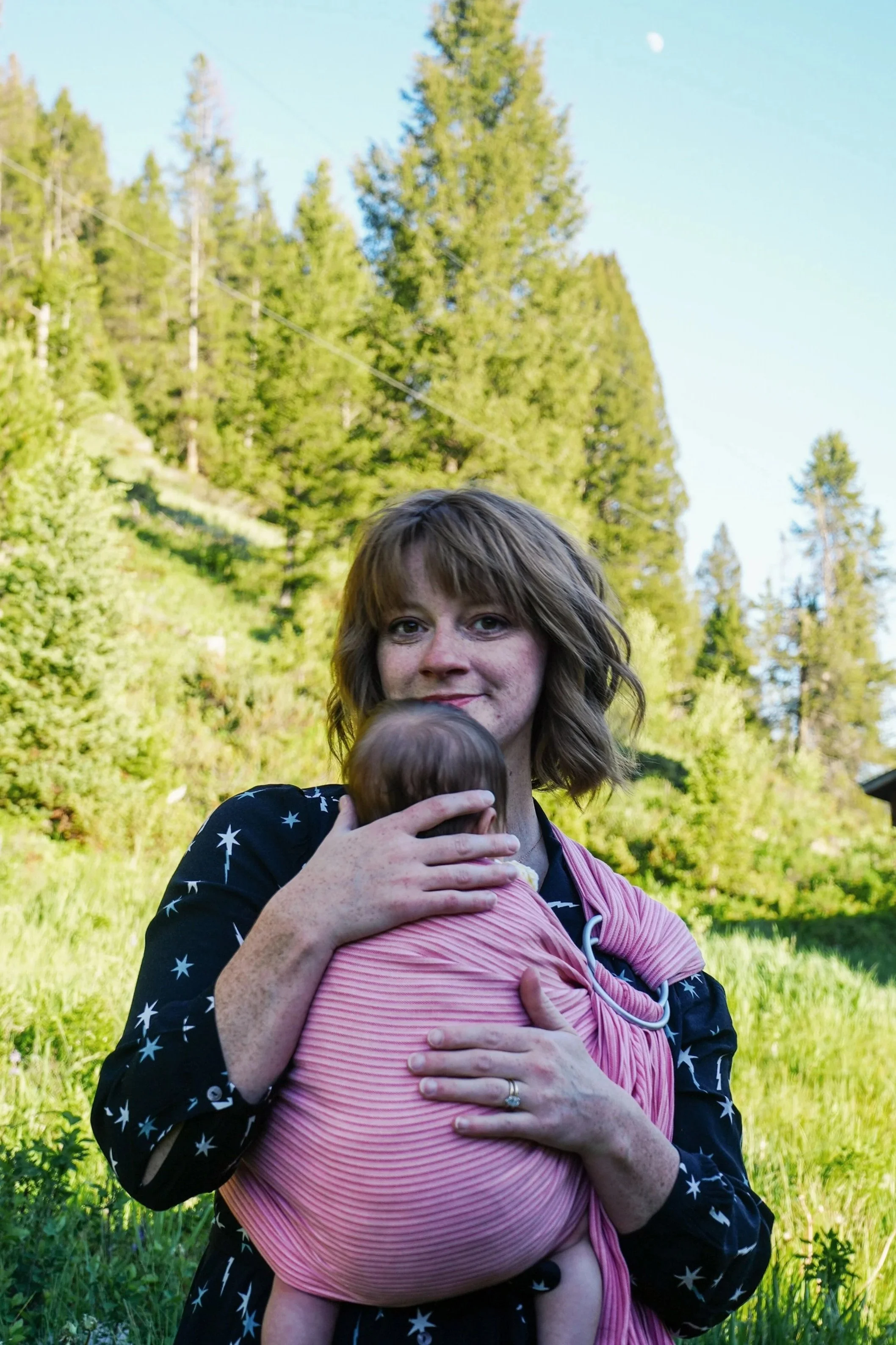 A woman with short, wavy light brown hair holding a small child wrapped in a pink blanket during daytime outdoors with green trees and grass in the background.