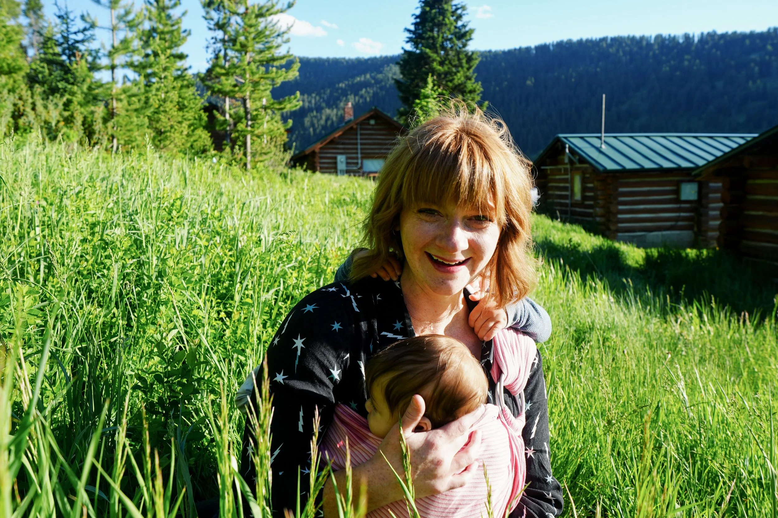 A woman holding two young children in a grassy field with tall green trees and wooden cabins in the background, under a blue sky.