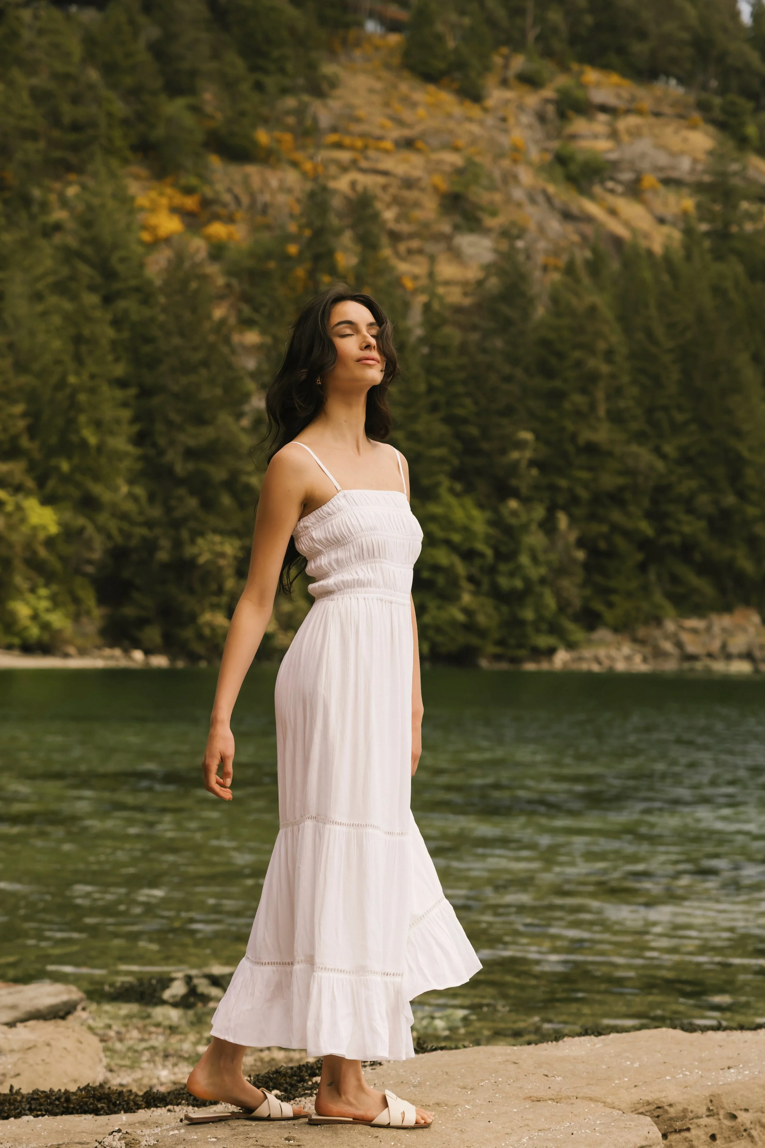 A woman wearing a white dress and sandals stands near a lake with a forest and mountains in the background.