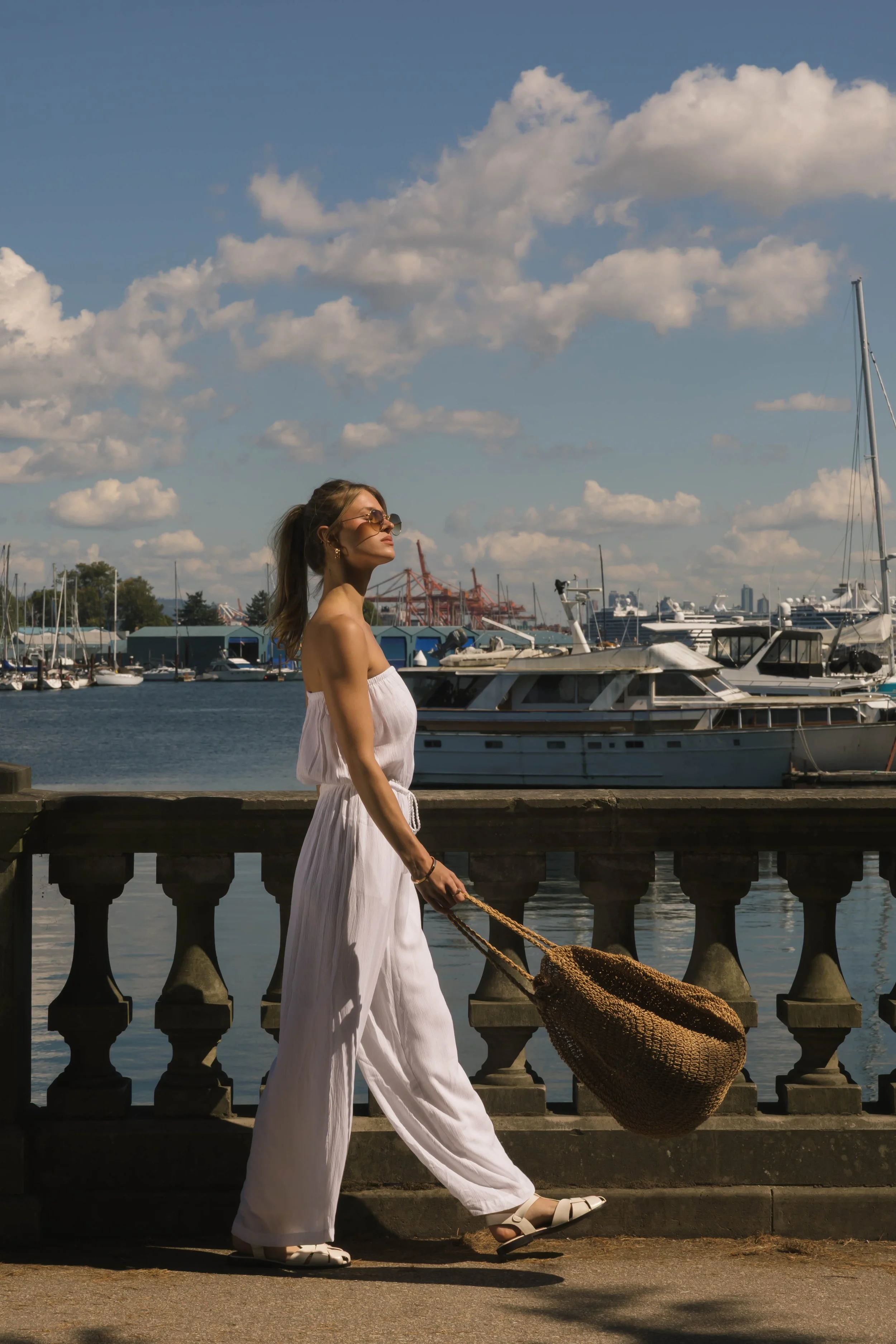 A woman in a white strapless jumpsuit and sunglasses walks along a waterfront, carrying a large woven bag. The background features boats docked in a marina under a partly cloudy sky.