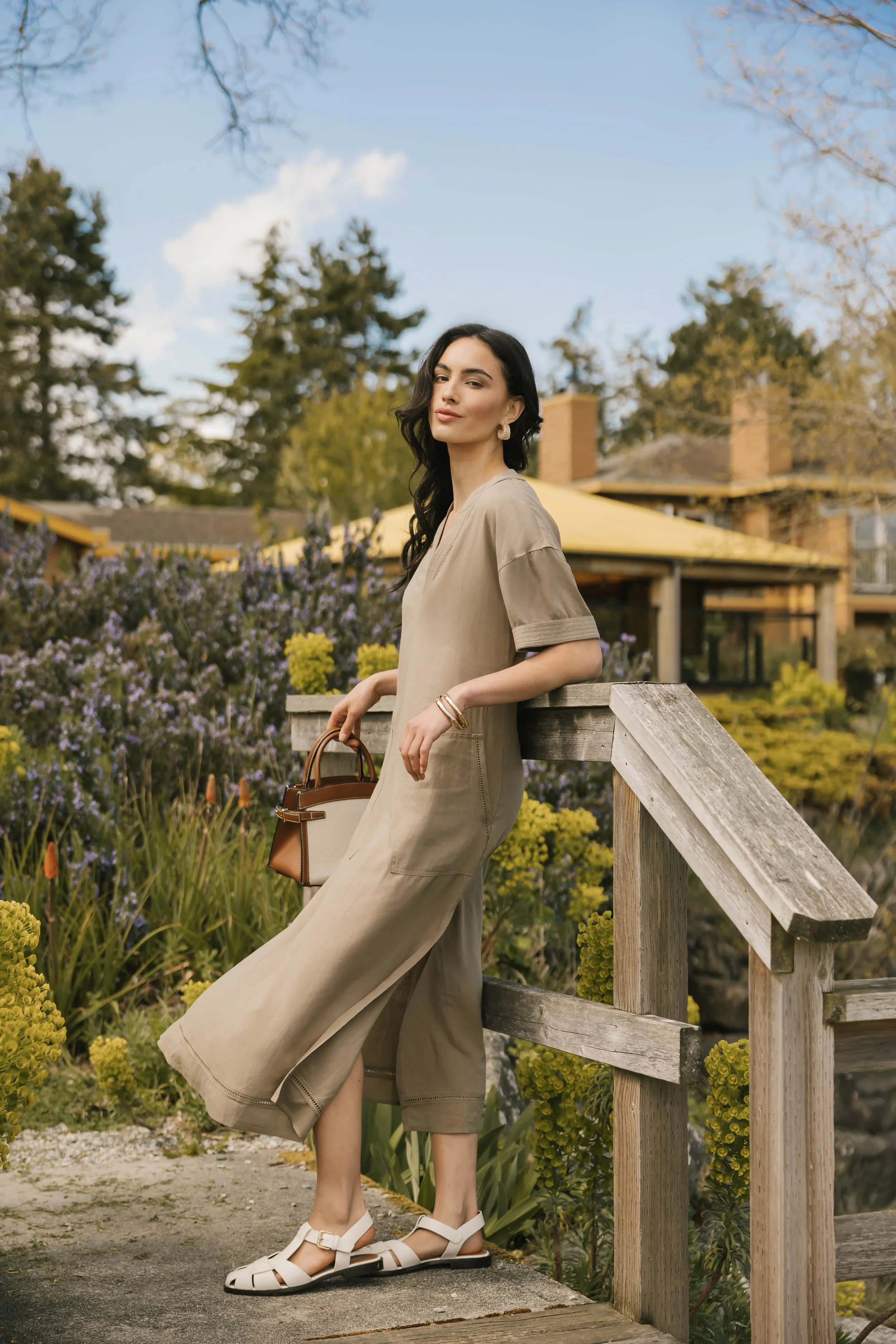 A woman in a beige dress holding a handbag, leaning against a wooden railing in a garden with purple flowers and greenery, with a house and trees in the background on a clear day.