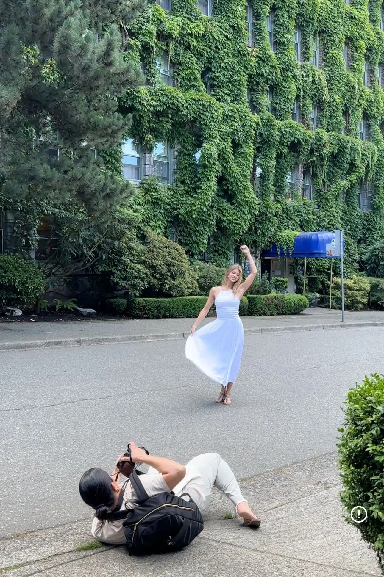 A woman in a white dress dances happily on an empty street while a photographer on the ground takes her picture, with green trees and a building with ivy in the background.