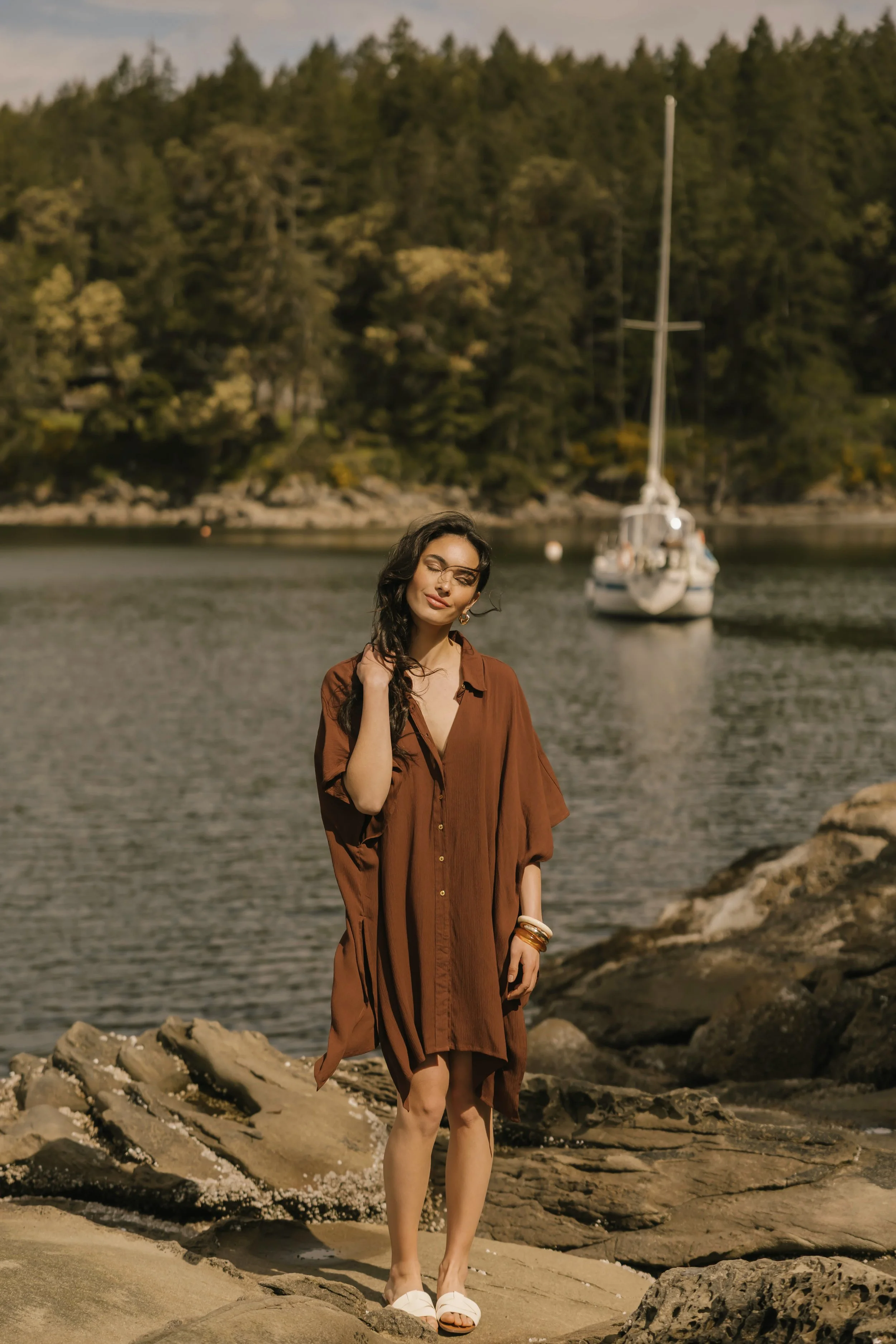 A woman in a brown dress standing on rocks by the water with a sailboat in the background and forested mountains beyond.