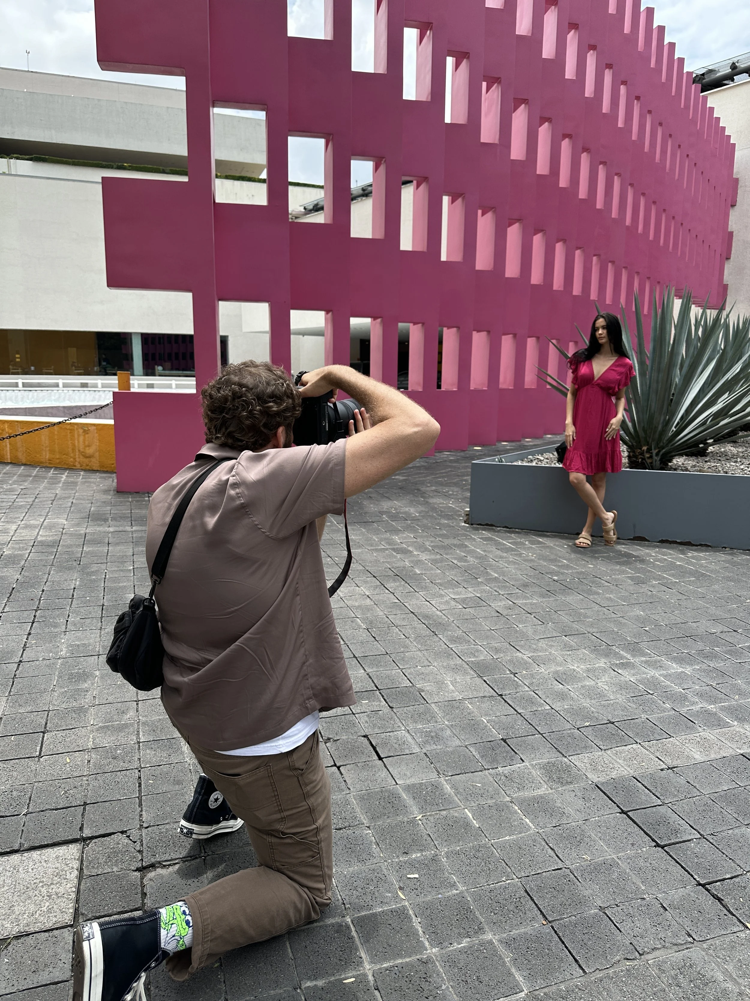 A man is taking a photograph of a woman in a pink dress outside near a large pink and gray architectural installation with a geometric pattern, in an outdoor urban setting.