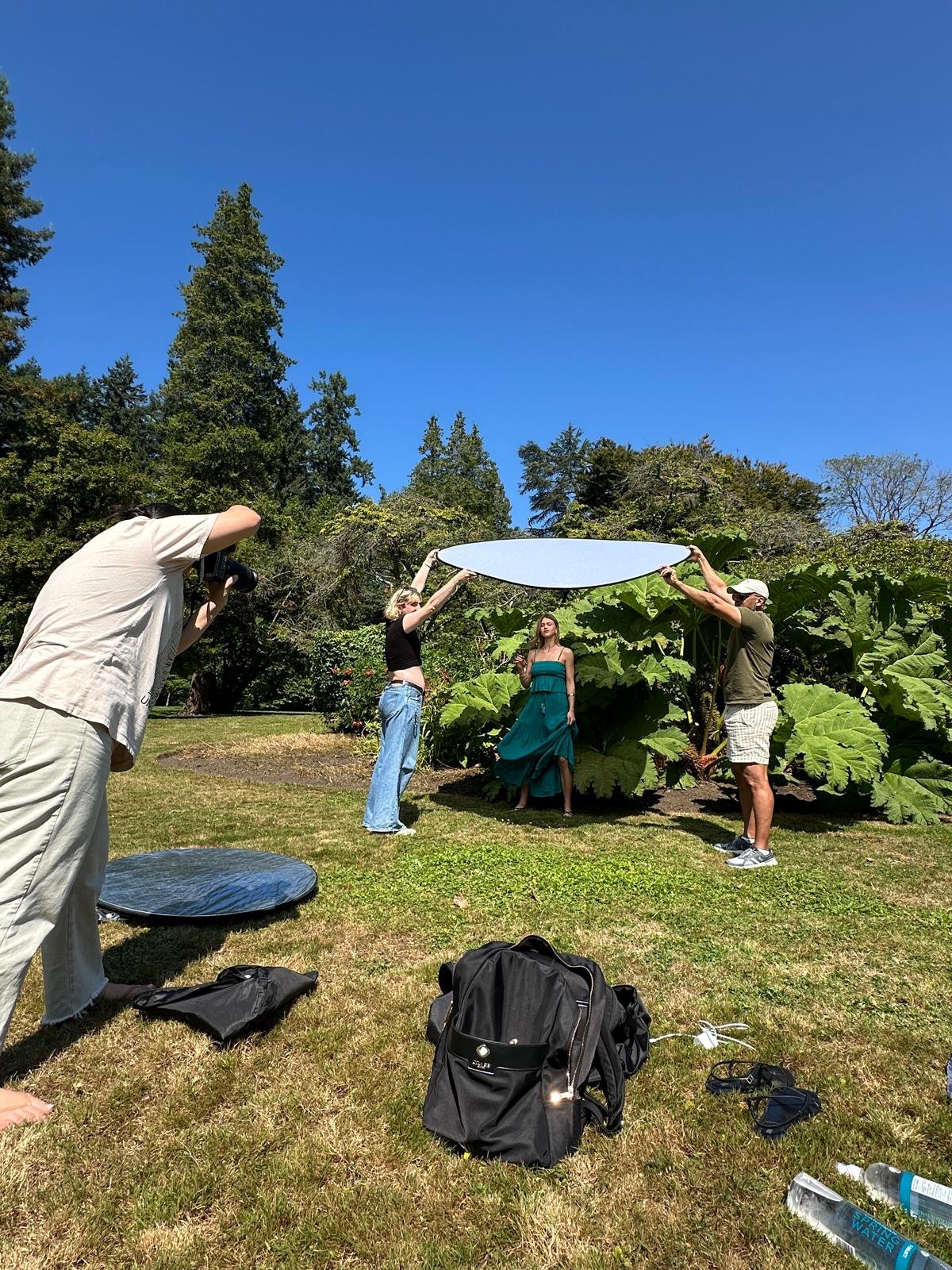 A woman in a teal dress is being photographed outdoors in a garden with large green leaves, while two individuals hold a circular reflector above her. A photographer is taking the picture, and various equipment and bags are on the ground.