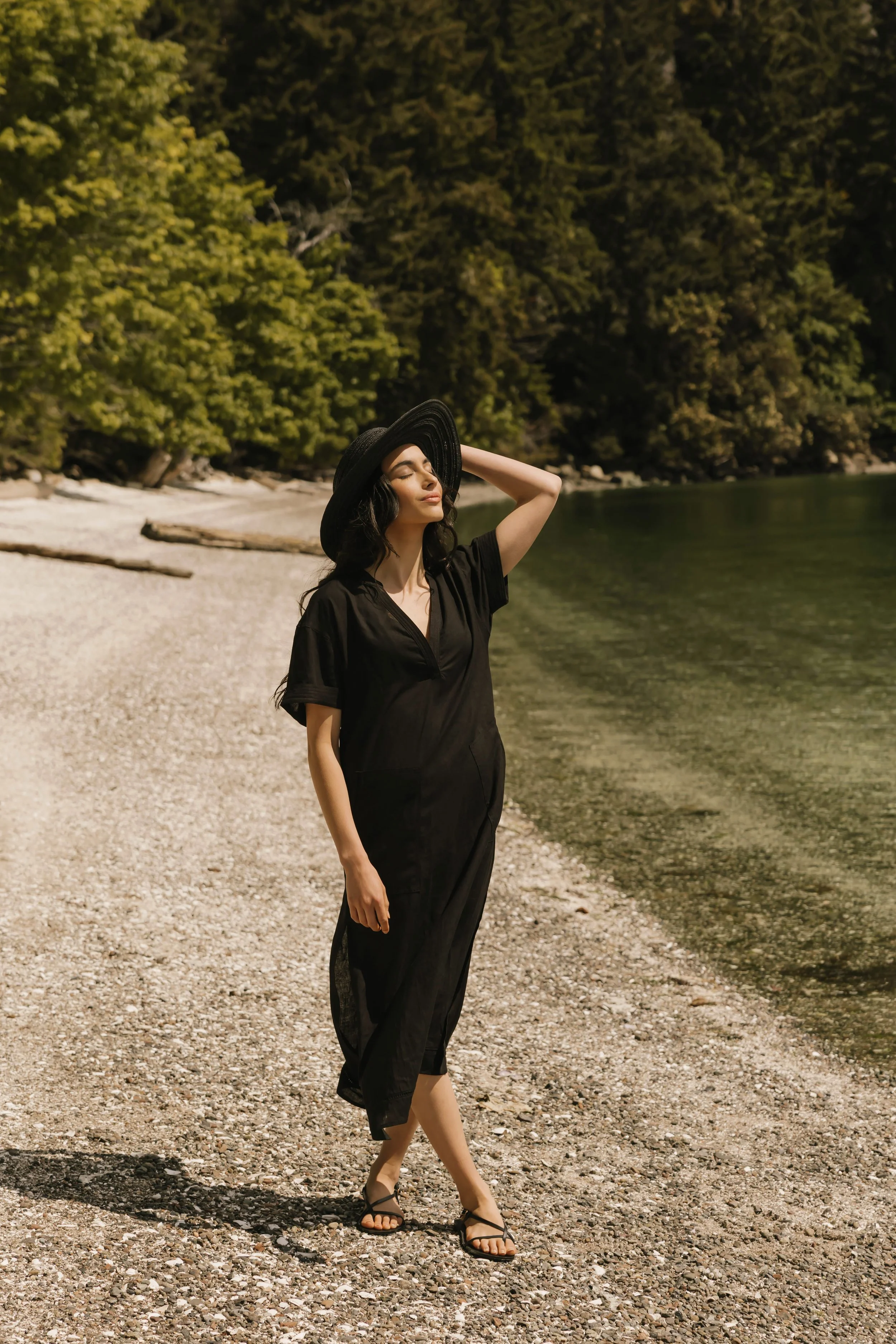 A woman in a black dress and black hat standing on a pebbled beach near the water, with trees in the background.