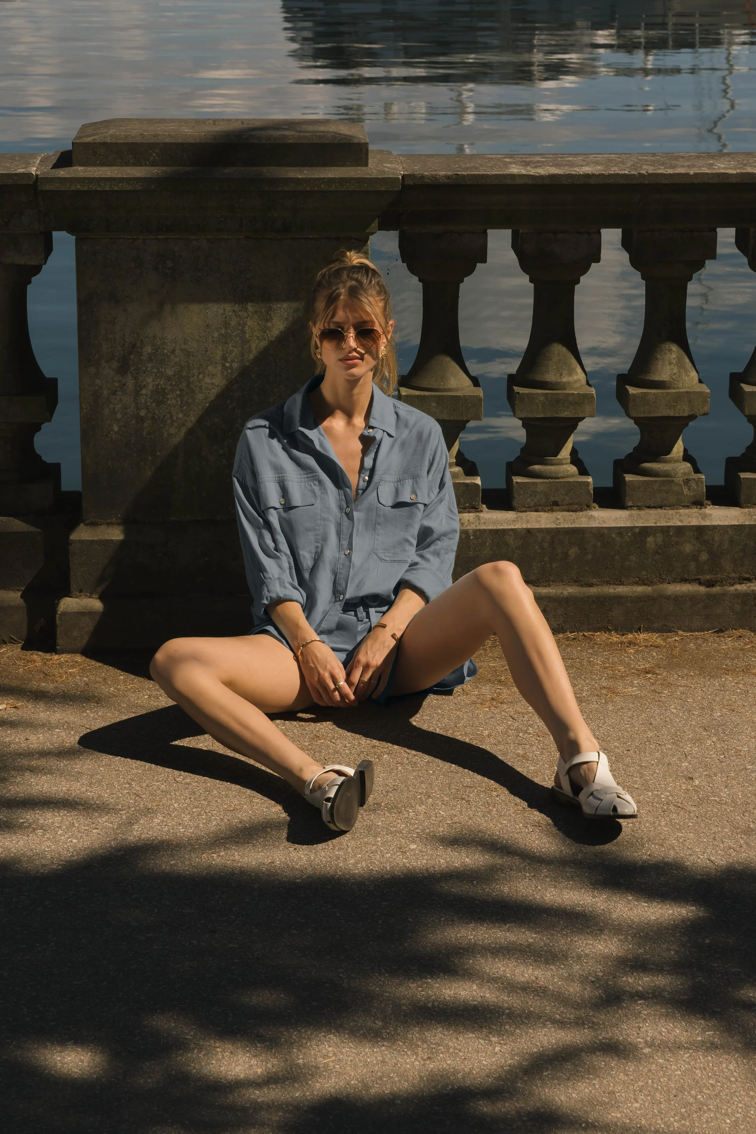 A young woman is sitting on the ground near a stone railing by a body of water, wearing sunglasses, an oversized blue button-up shirt, and white shoes. She is leaning against the railing in sunlight, with shadows cast from nearby trees.