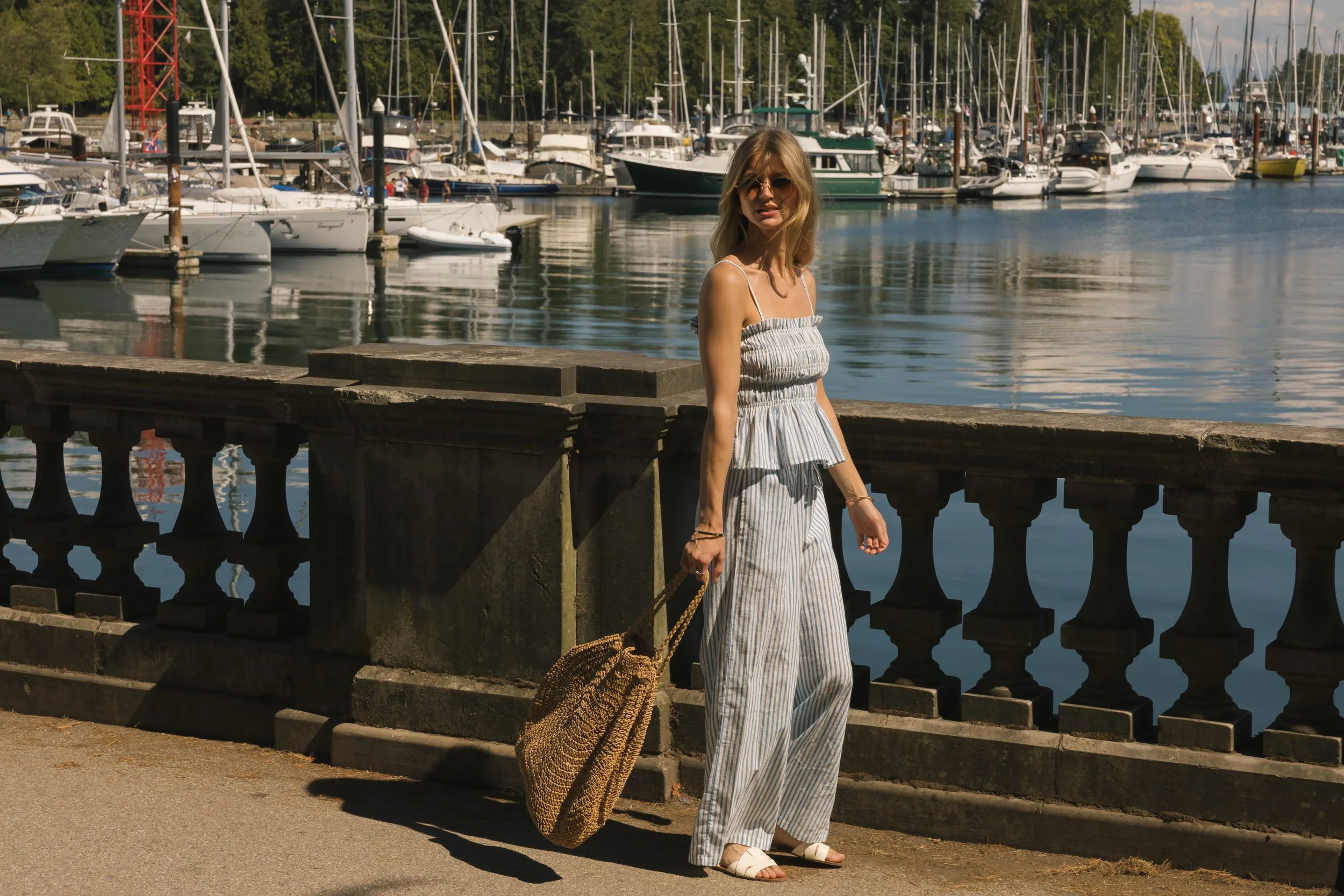 A woman with blonde hair wearing sunglasses, a white and gray striped sleeveless top, and matching wide-leg pants, walks along a waterfront promenade with boats and sailboats docked in the background.
