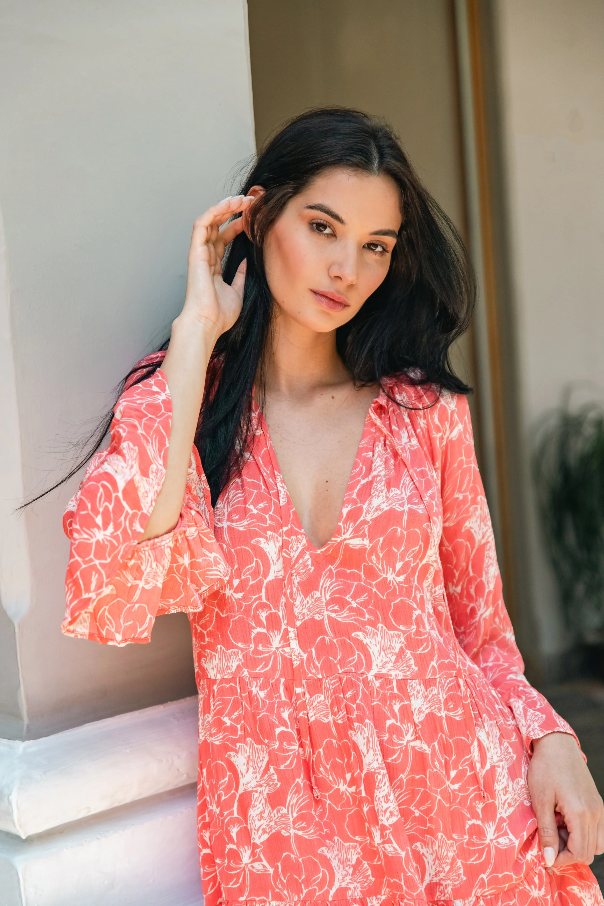 A woman with long black hair in a coral-colored floral dress leaning against a wall indoors.