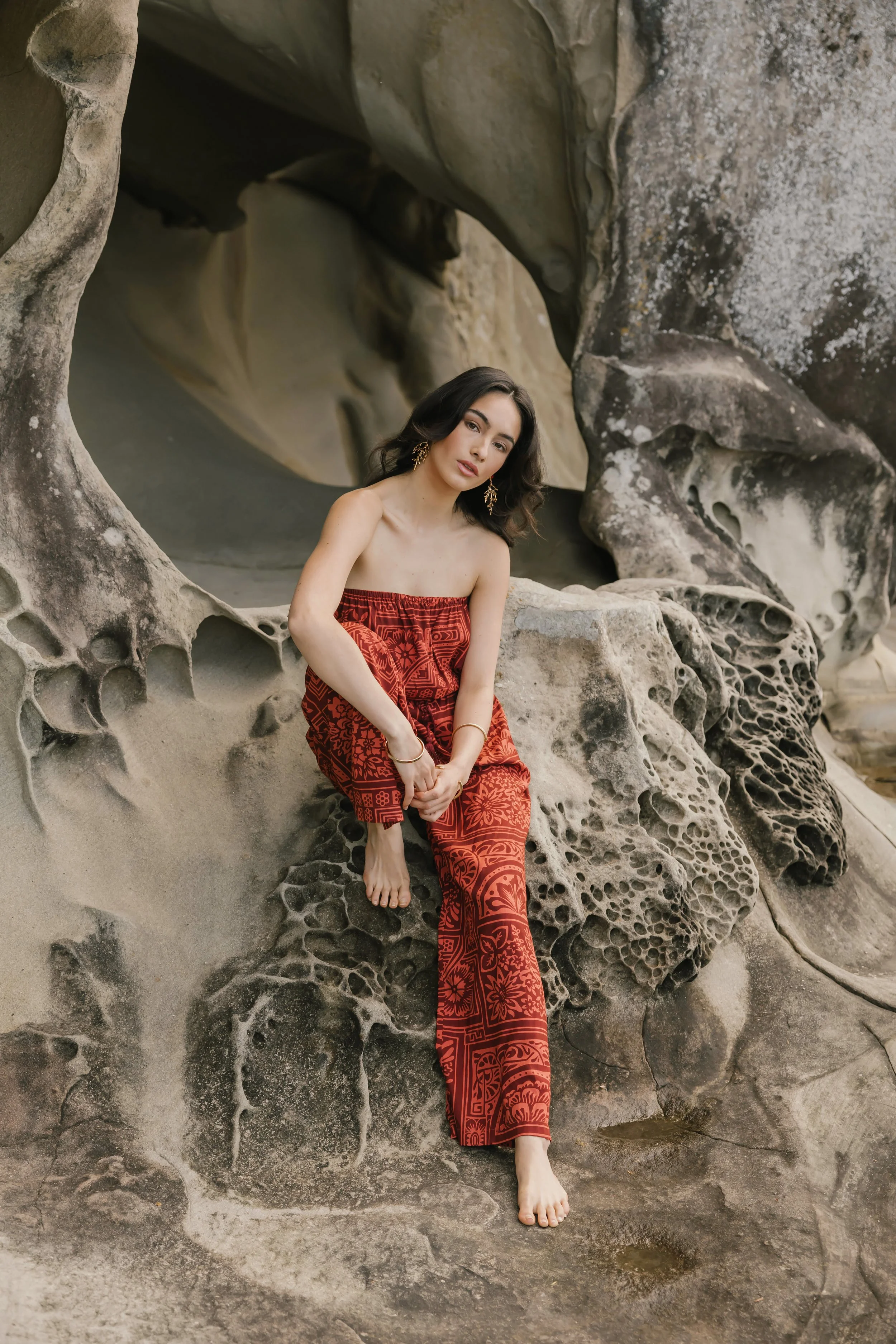 A woman sitting on a large, textured rock formation with intricate patterns, wearing a red strapless dress and gold jewelry, posed against a natural rocky background.