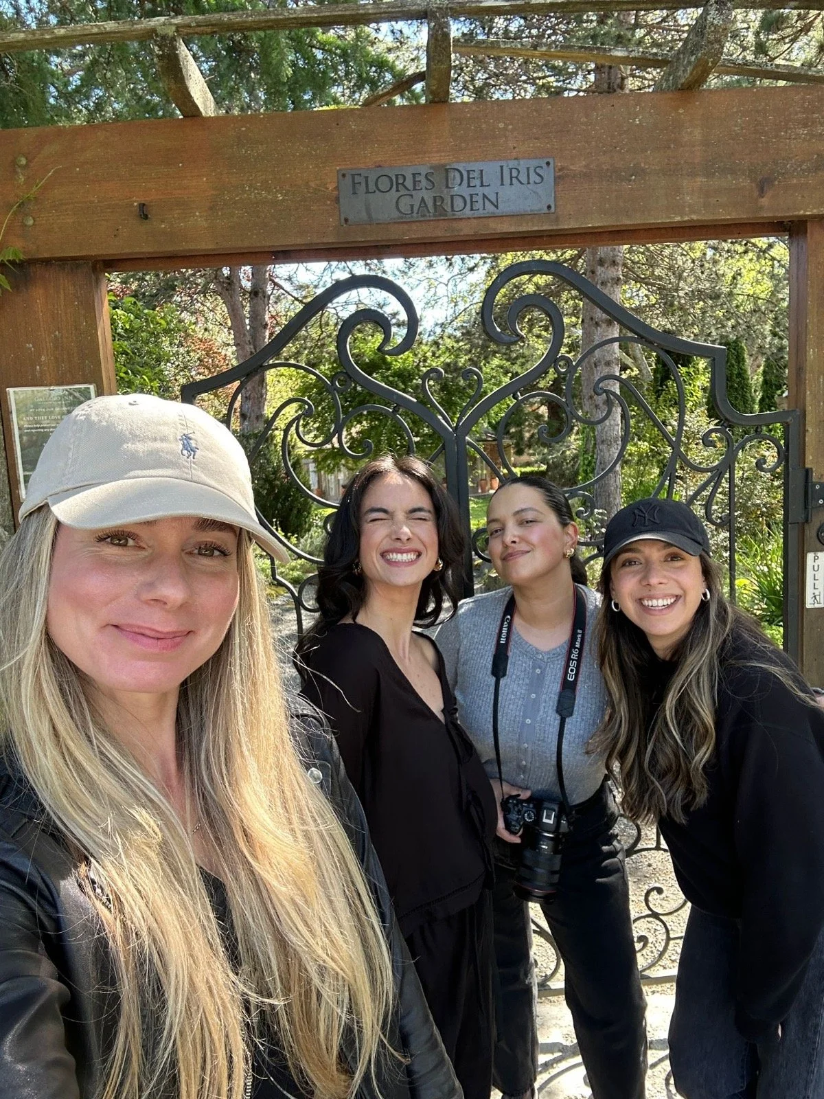Four women smiling at the camera in front of a garden gate, with a sign that reads 'Flores del Iris Garden'.
