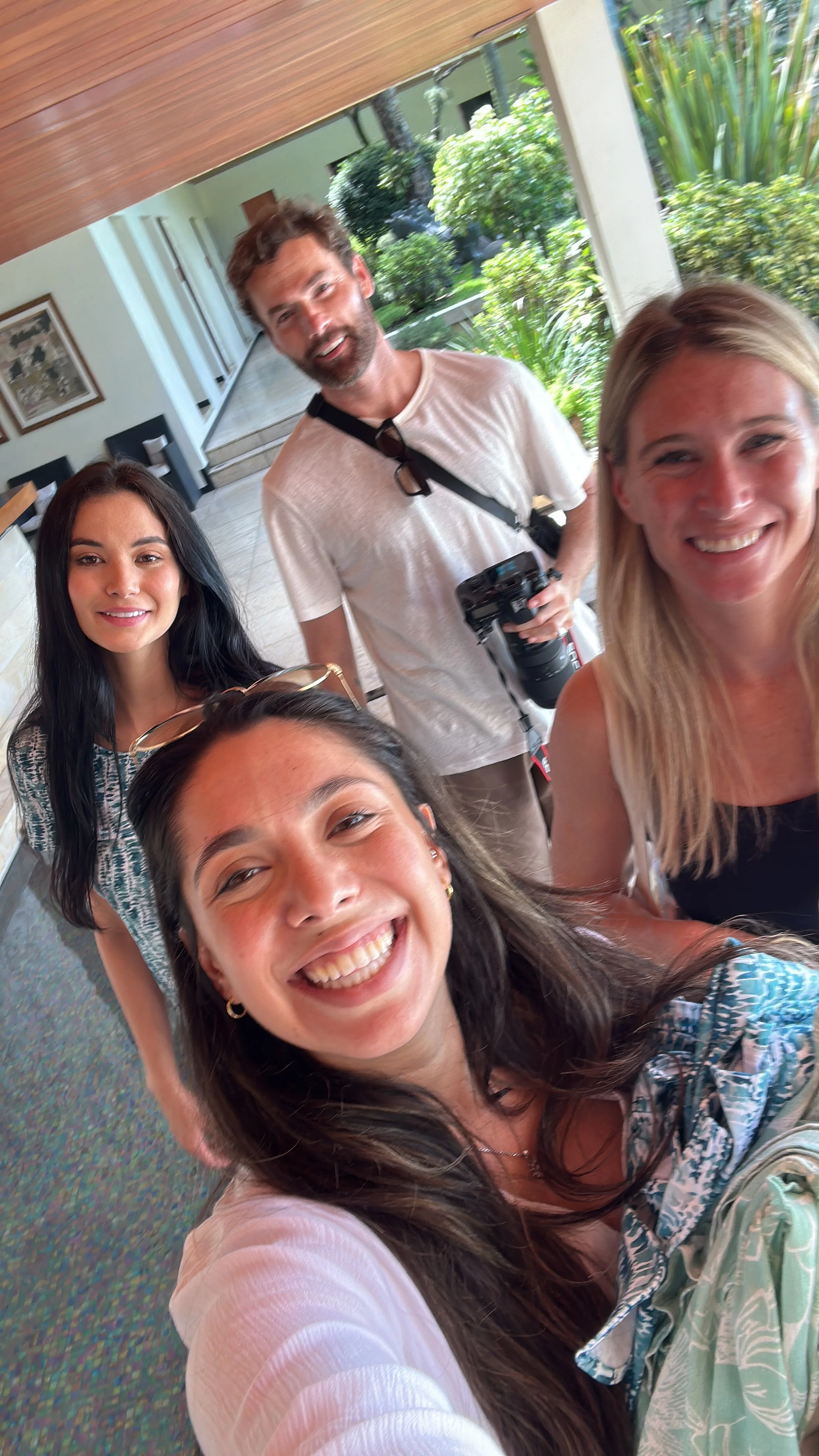 Group of five smiling people taking a selfie indoors with a view of a lush green garden outside.