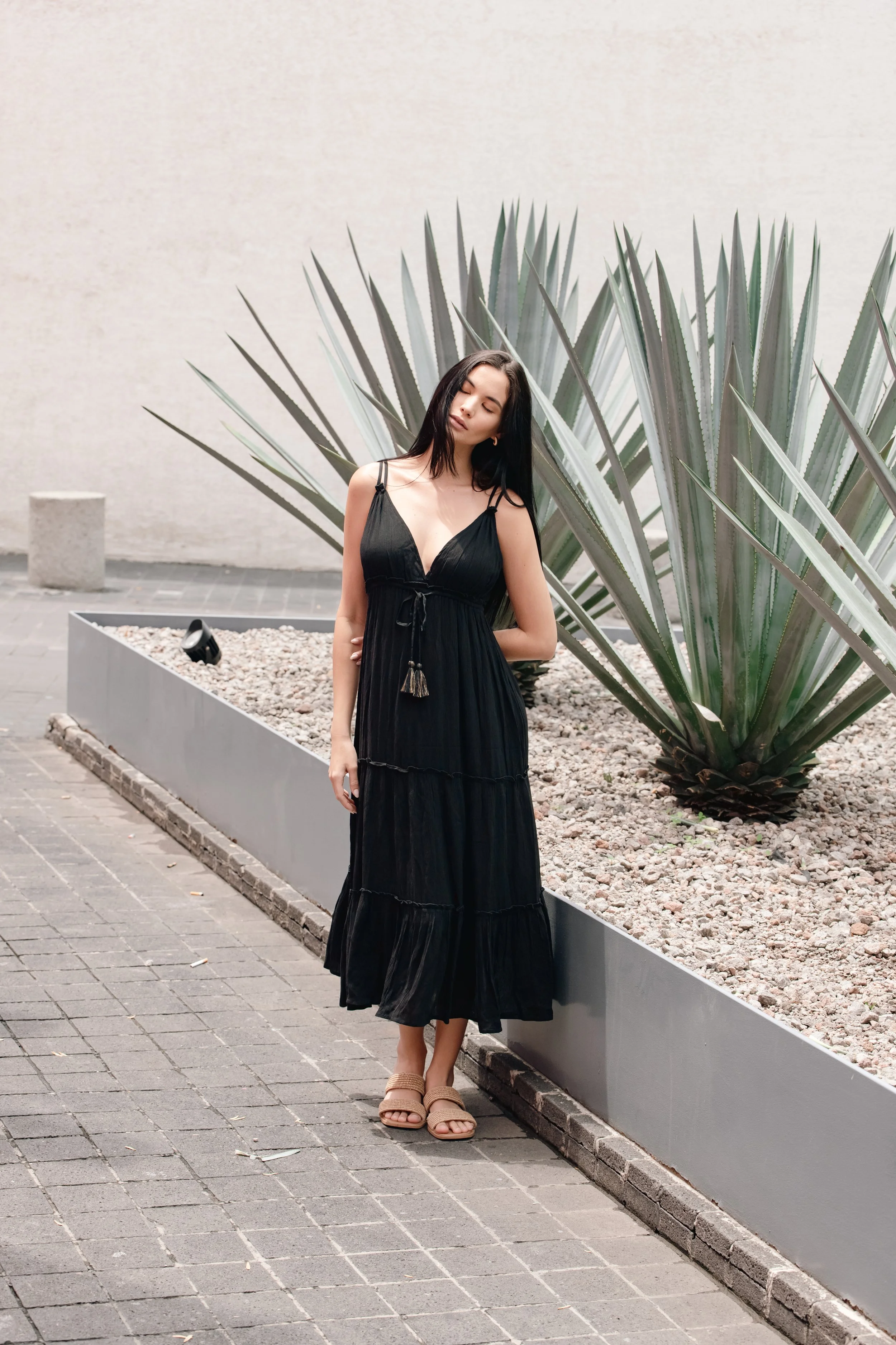 A woman in a black summer dress standing outdoors next to a large agave plant with her eyes closed.