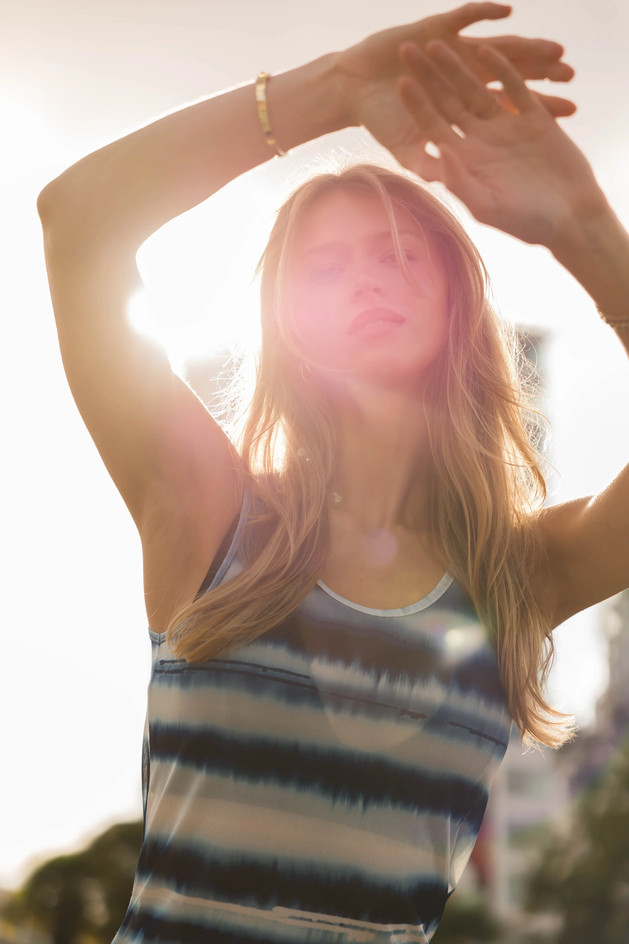 A young woman with long hair stands outdoors during sunset, with her arms raised above her head, casting a glow around her.