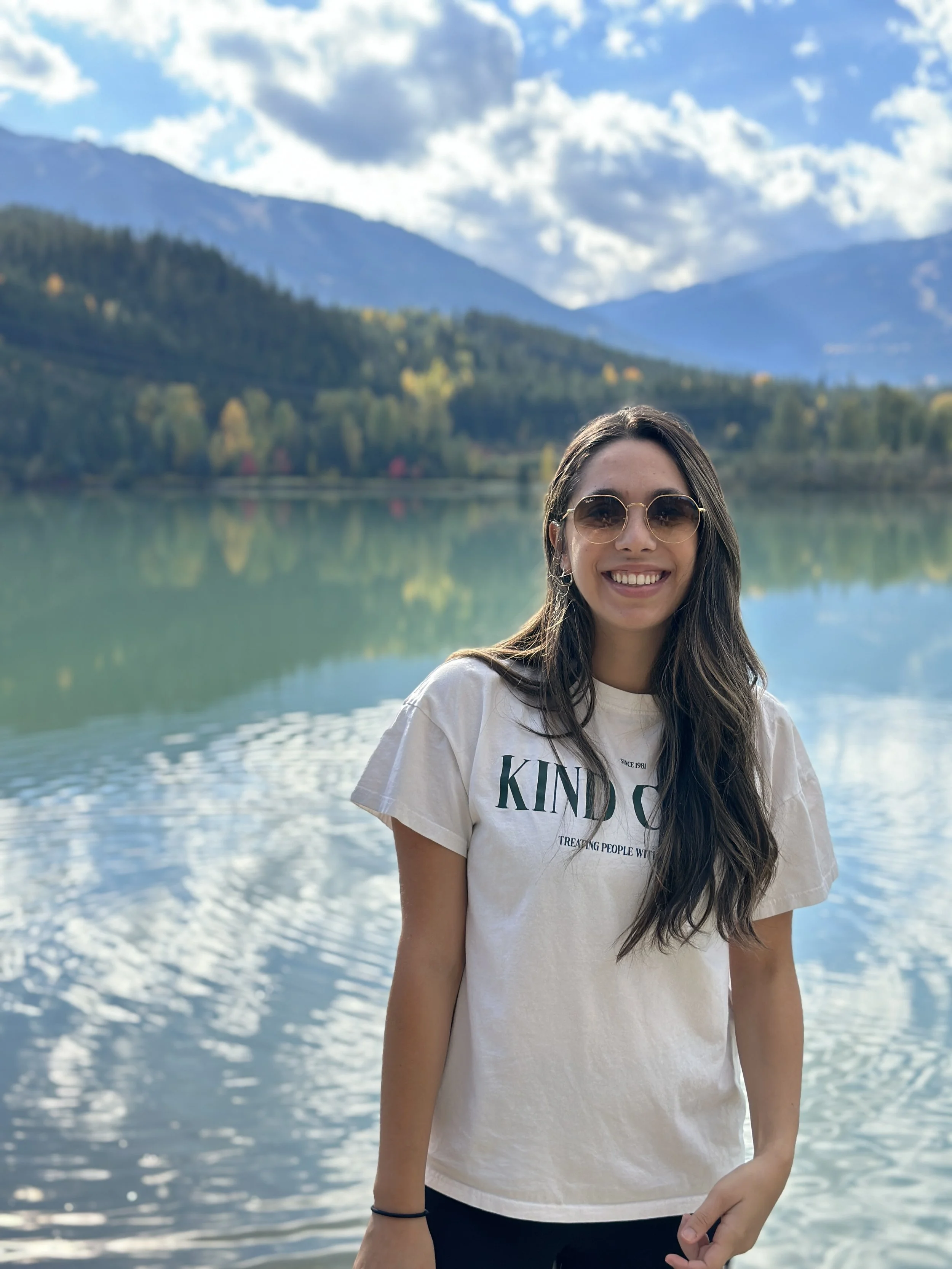 A woman with long brown hair and sunglasses smiling in front of a body of water with mountains and trees in the background.